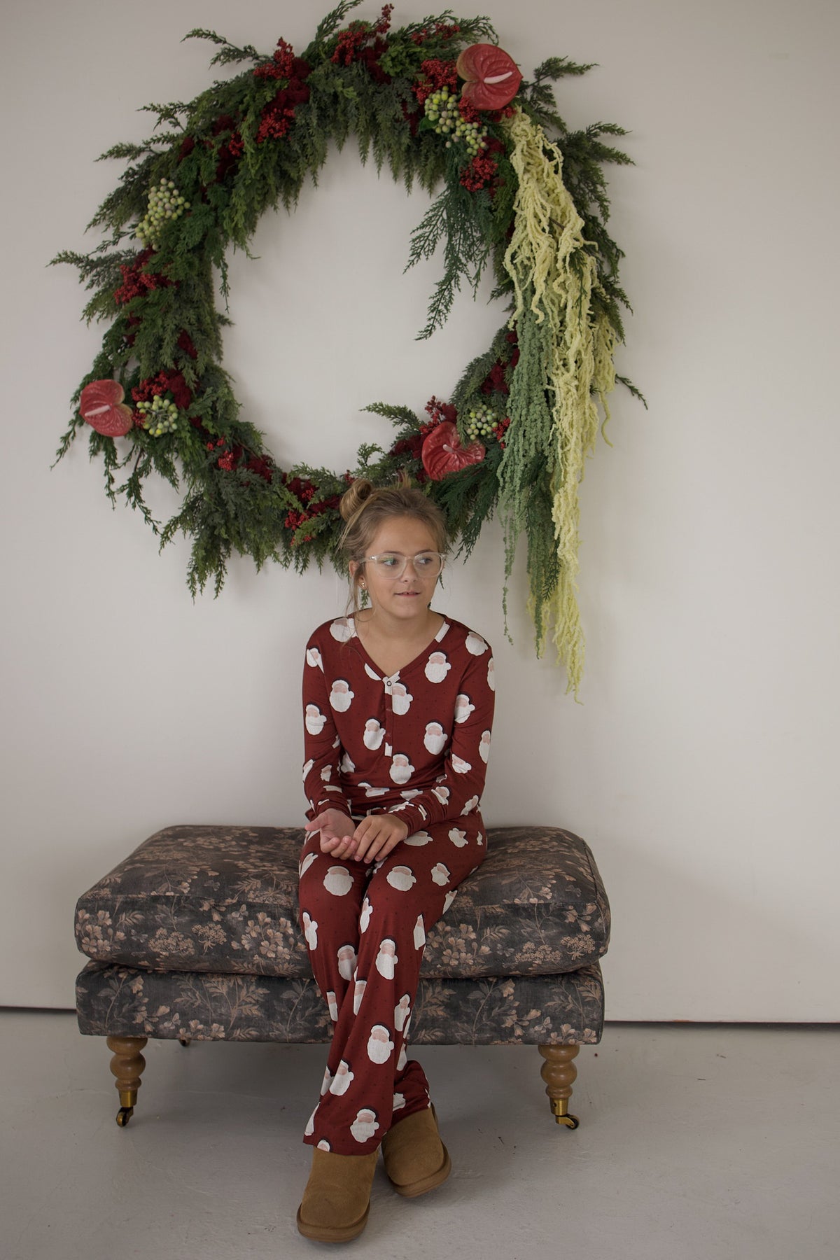 A child wearing a red long-sleeve Youth Flare Bamboo Pajamas | Vintage Santa set, sitting on a patterned ottoman in front of a large holiday wreath.