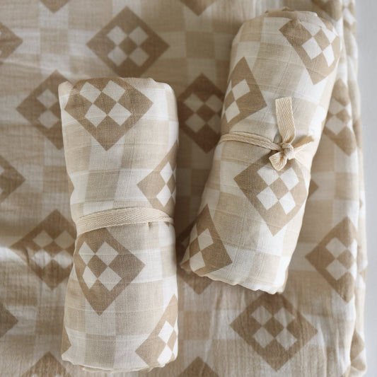 Two rolled forever french baby Muslin Swaddle | Cottage Quilt blankets with beige and white diamond designs, each tied with fabric bands, rest on a matching patterned background.