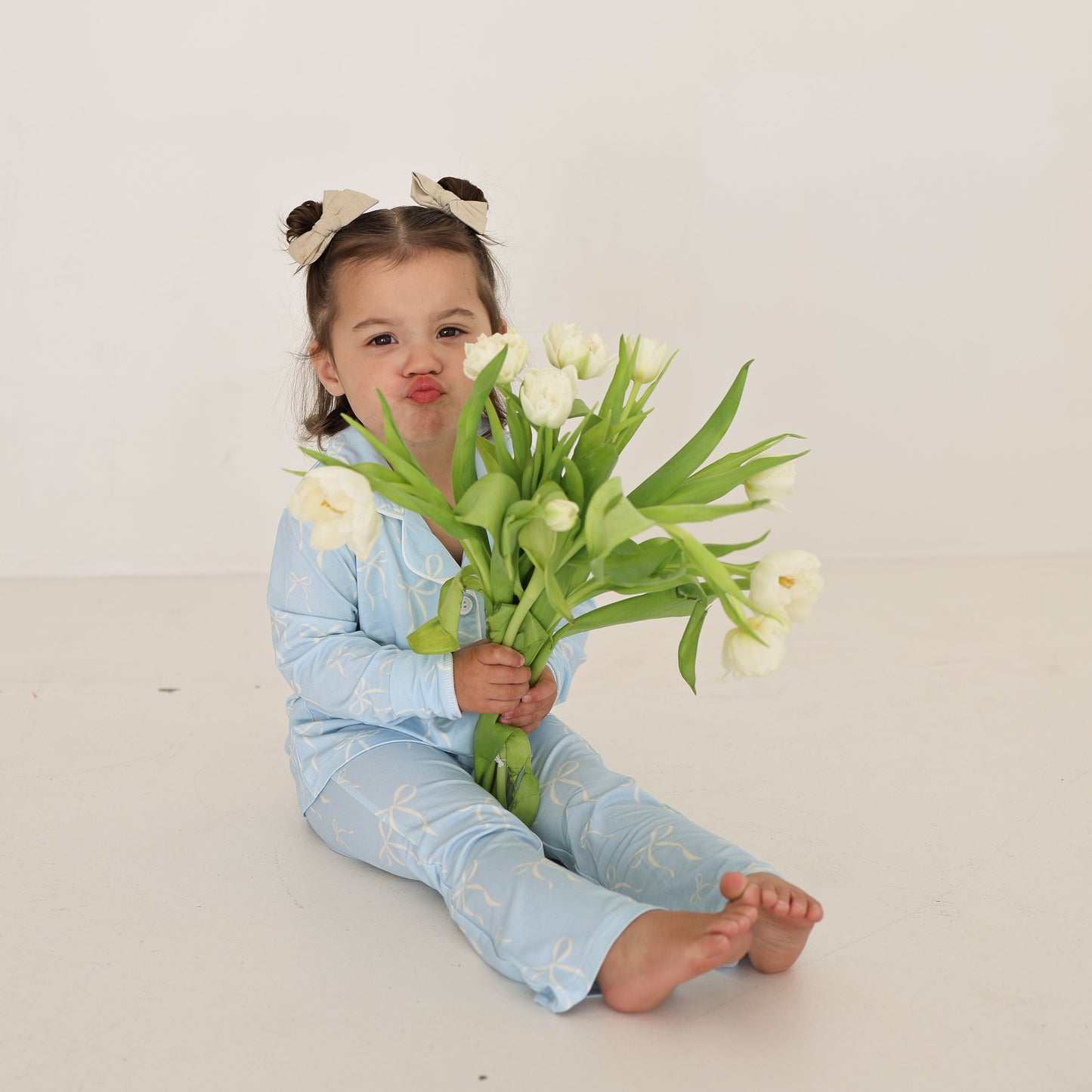 A young girl in a soft, hypo-allergenic forever french baby Girl’s Bamboo Button Up Lounge Set | Blue Bows sits barefoot with panda ear hair accessories, holding white tulips and pouting playfully against a plain white background.