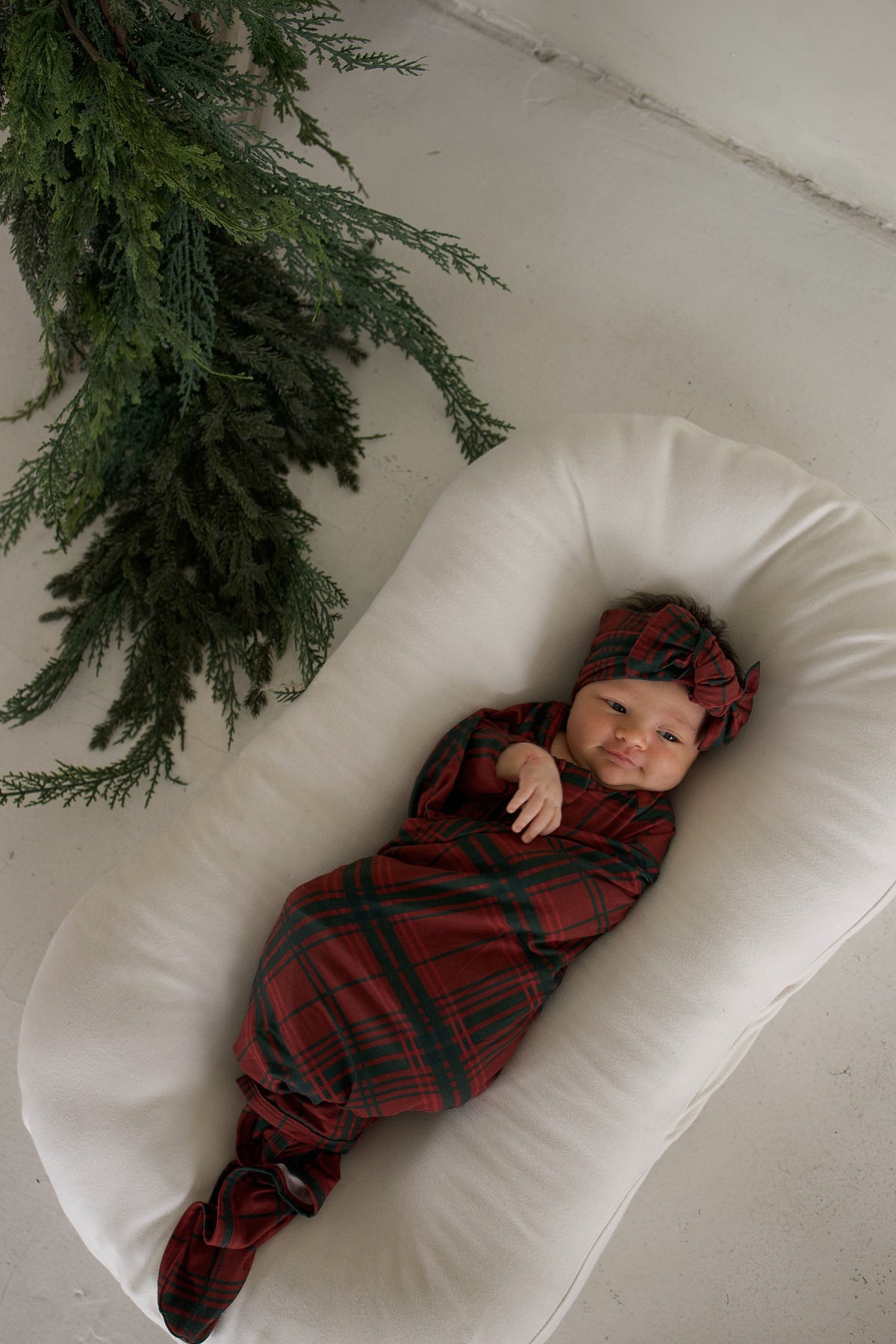 Newborn baby wrapped in a forever french baby Bamboo Swaddle | Christmas Plaid with a matching knotted headband, resting in a white baby lounger.