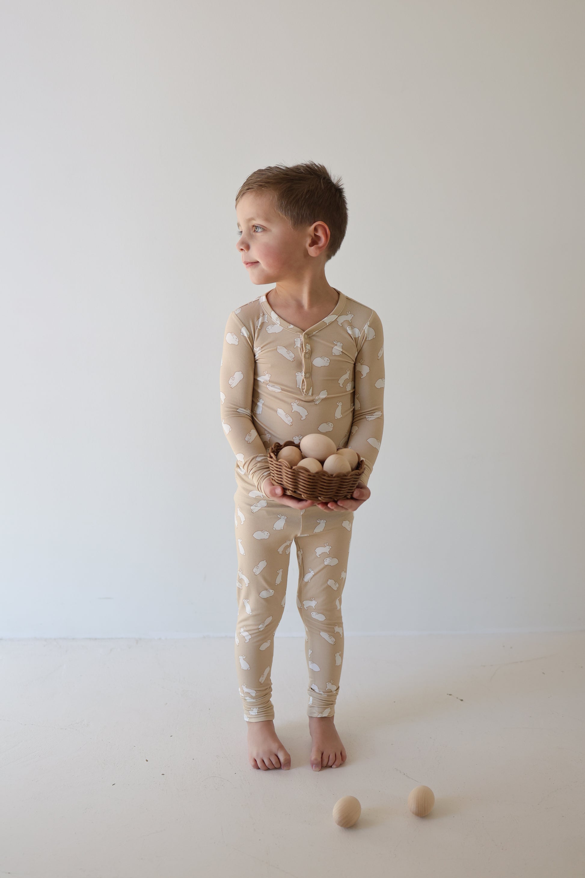 A young child in the forever french baby Bamboo Two Piece Pajamas | Bunnies, barefoot on a white floor, holds a bowl of wooden eggs. Three wooden eggs lie near their feet. The background is plain and light.