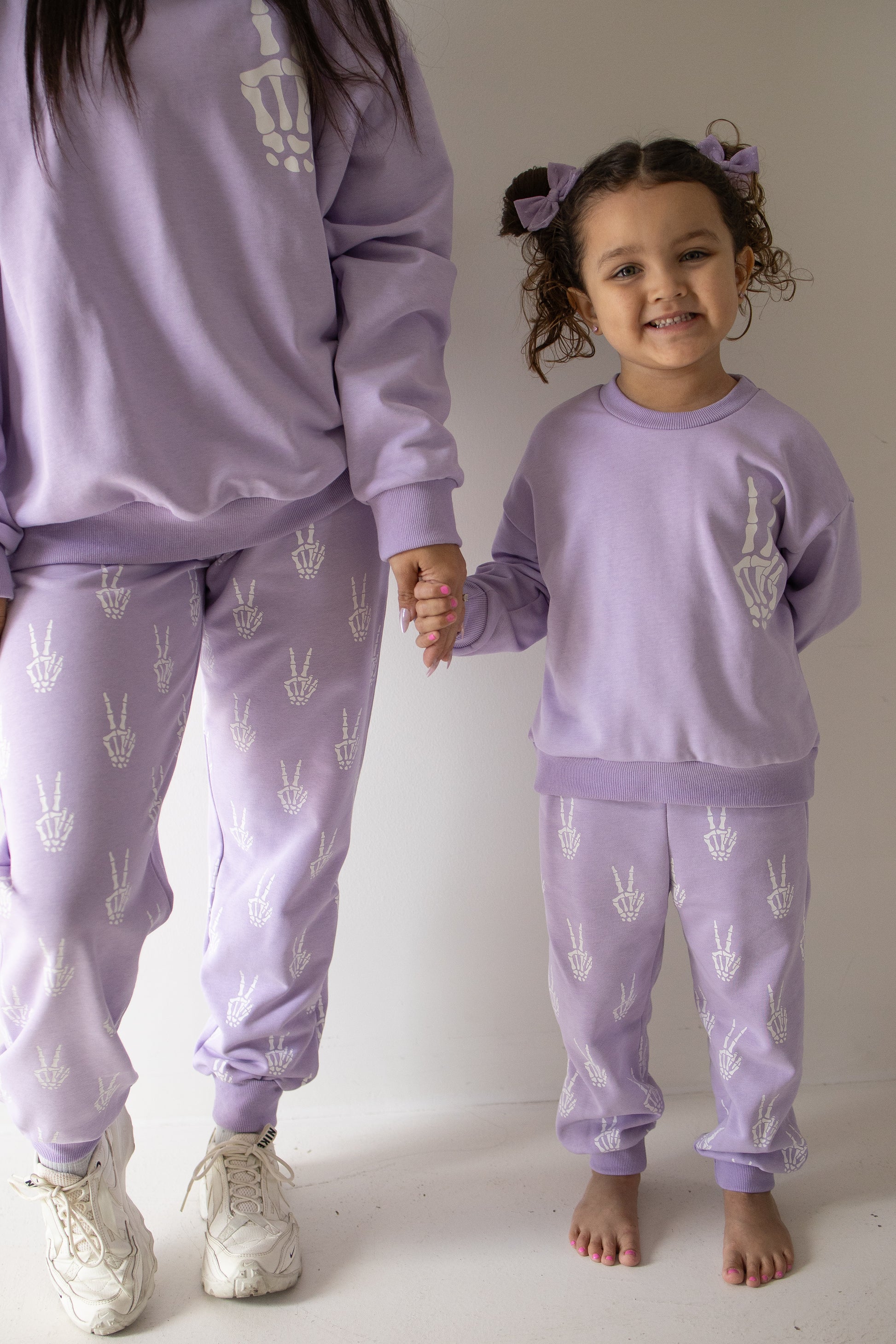 An adult and a child hold hands, both dressed in matching lavender “Enchanted Skelly” sweatsuits by forever french baby, featuring playful white skeleton hand designs. The child, in the Child Sweat Set | Enchanted Skelly, smiles and stands barefoot beside the adult, who wears white sneakers—a fun outfit for everyday adventures.