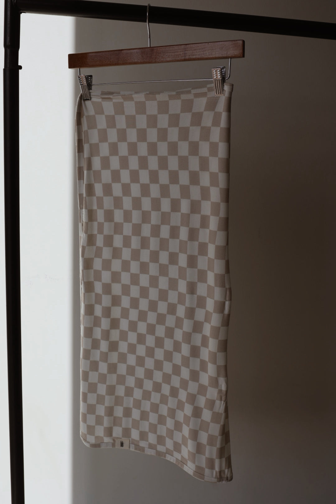 The forever french baby Bamboo Swaddle | Checkerboard, in beige and white checkered fabric, hangs from a wooden hanger on a black clothing rack against a softly lit plain background, echoing gentle comfort and breathability.