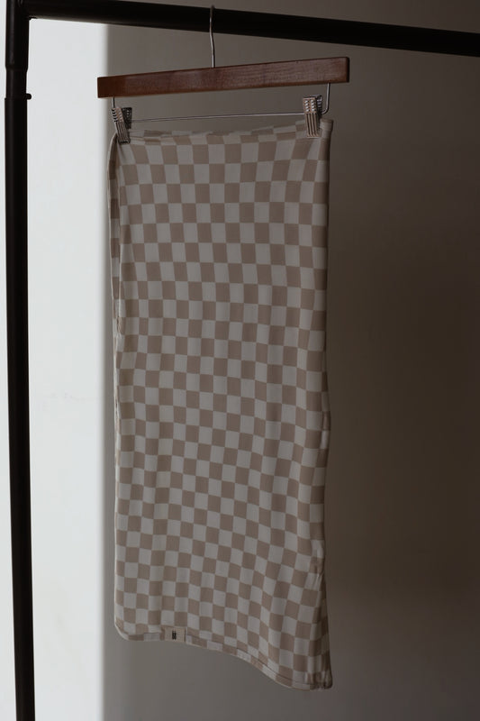 The forever french baby Bamboo Swaddle | Checkerboard, in beige and white checkered fabric, hangs from a wooden hanger on a black clothing rack against a softly lit plain background, echoing gentle comfort and breathability.