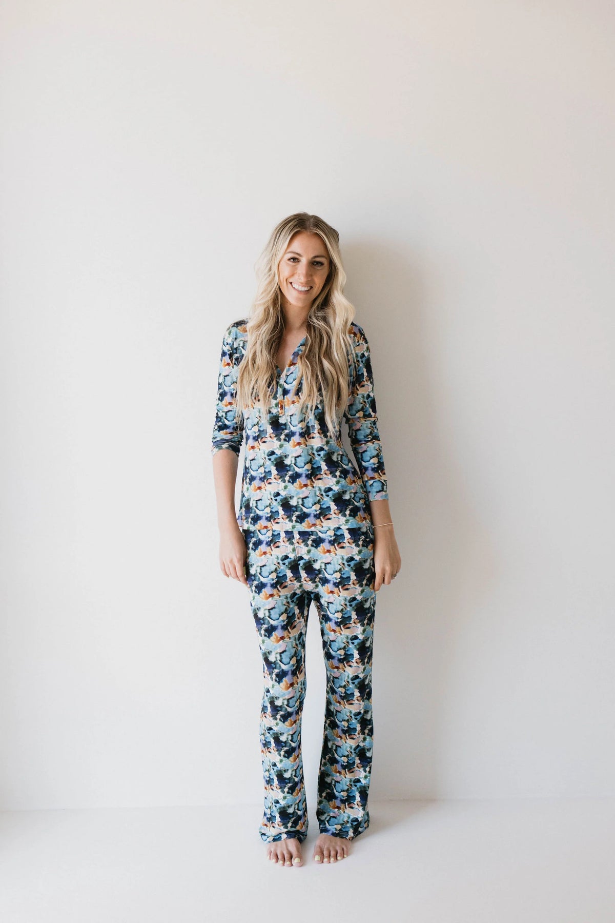 A person with long blonde hair is standing barefoot against a plain white wall, smiling at the camera while wearing the forever french baby Bamboo Two Piece Pajamas in the Charli Print.