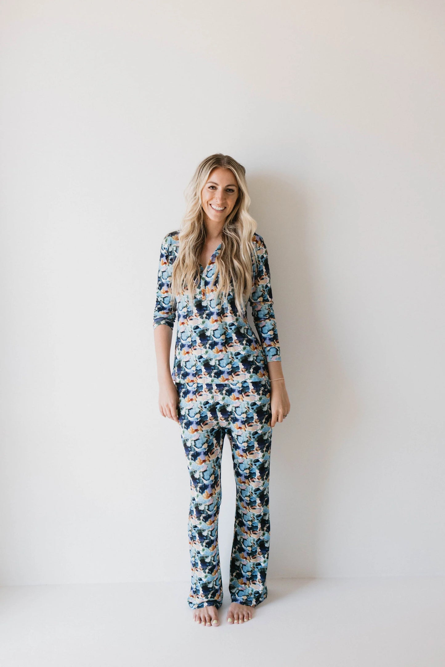 A smiling person with long, wavy blonde hair wears the forever french baby Bamboo Two Piece Pajamas in the Charli Print—an autism awareness-inspired, colorful floral set—and stands barefoot against a plain white wall.