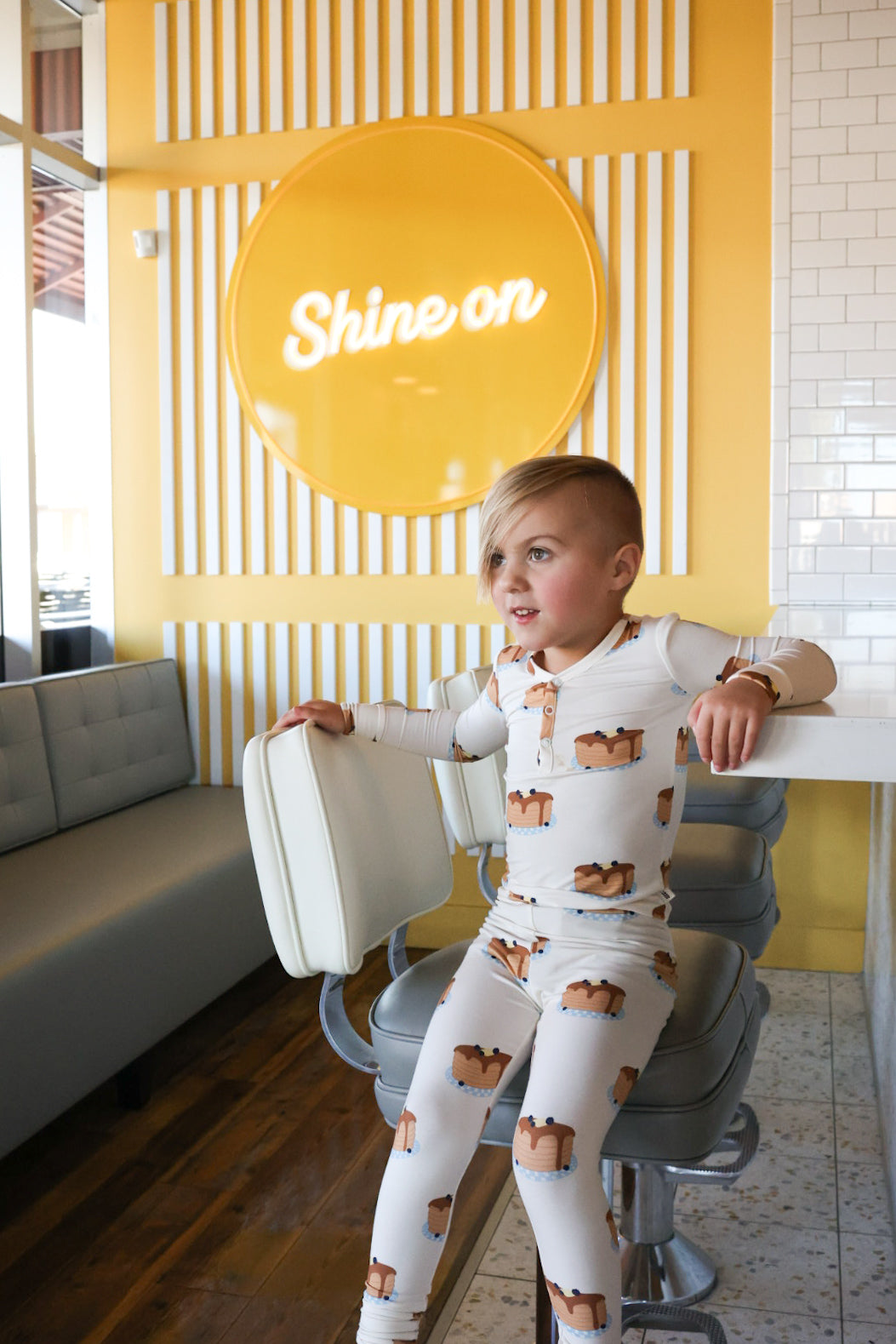 A young child with short blond hair sits on a white retro diner stool, wearing forever french baby Bamboo Two Piece Pajamas | Sunday Stack. Behind them is a yellow wall with white stripes and a "Shine on" sign.