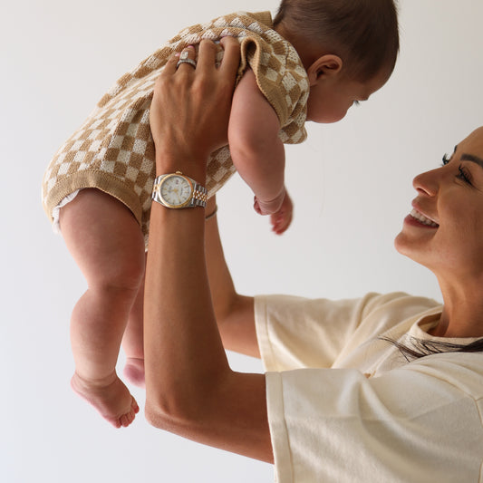 A smiling adult in a white shirt lifts a happy baby wearing the forever french baby Knit Romper | Latte Check, featuring a brown and white checkerboard print, both against a plain white background.