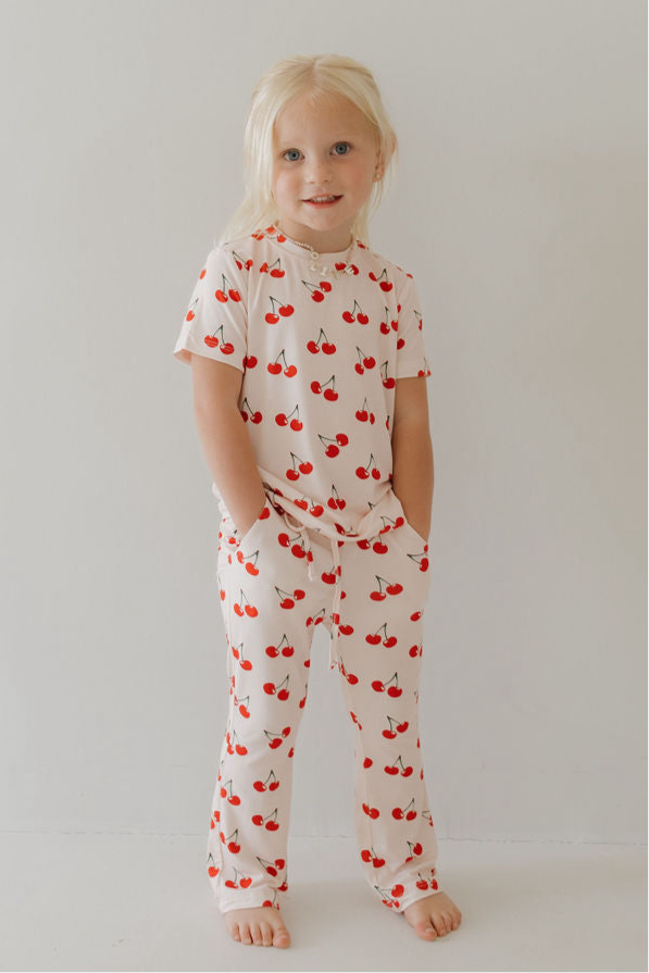 A young child with long blonde hair stands barefoot in the forever french baby Toddler Short Sleeve Bamboo Lounge Set | Cherry Orchard, featuring a white bamboo fabric with a red cherry pattern, hands in pockets, enjoying its breathable comfort.