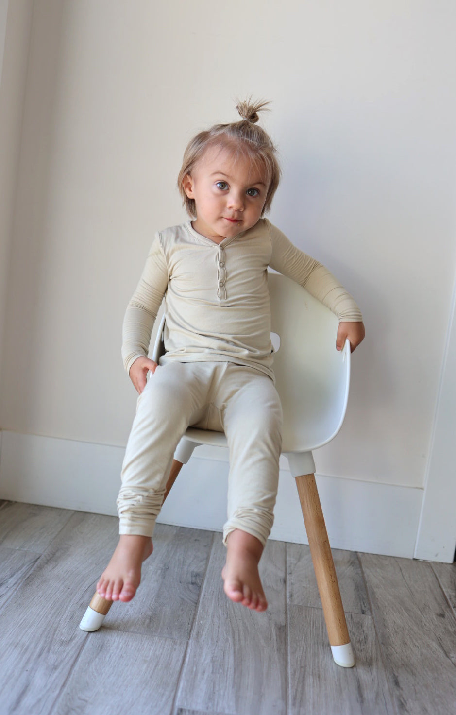 A young child with light hair in a top knot sits on a white chair with wooden legs, wearing the Beige Long-Sleeved Bamboo Two Piece Pajamas named "Limestone" from forever french baby. The child gazes slightly to the side with a curious expression against a minimalist backdrop of a white wall and wooden floor.