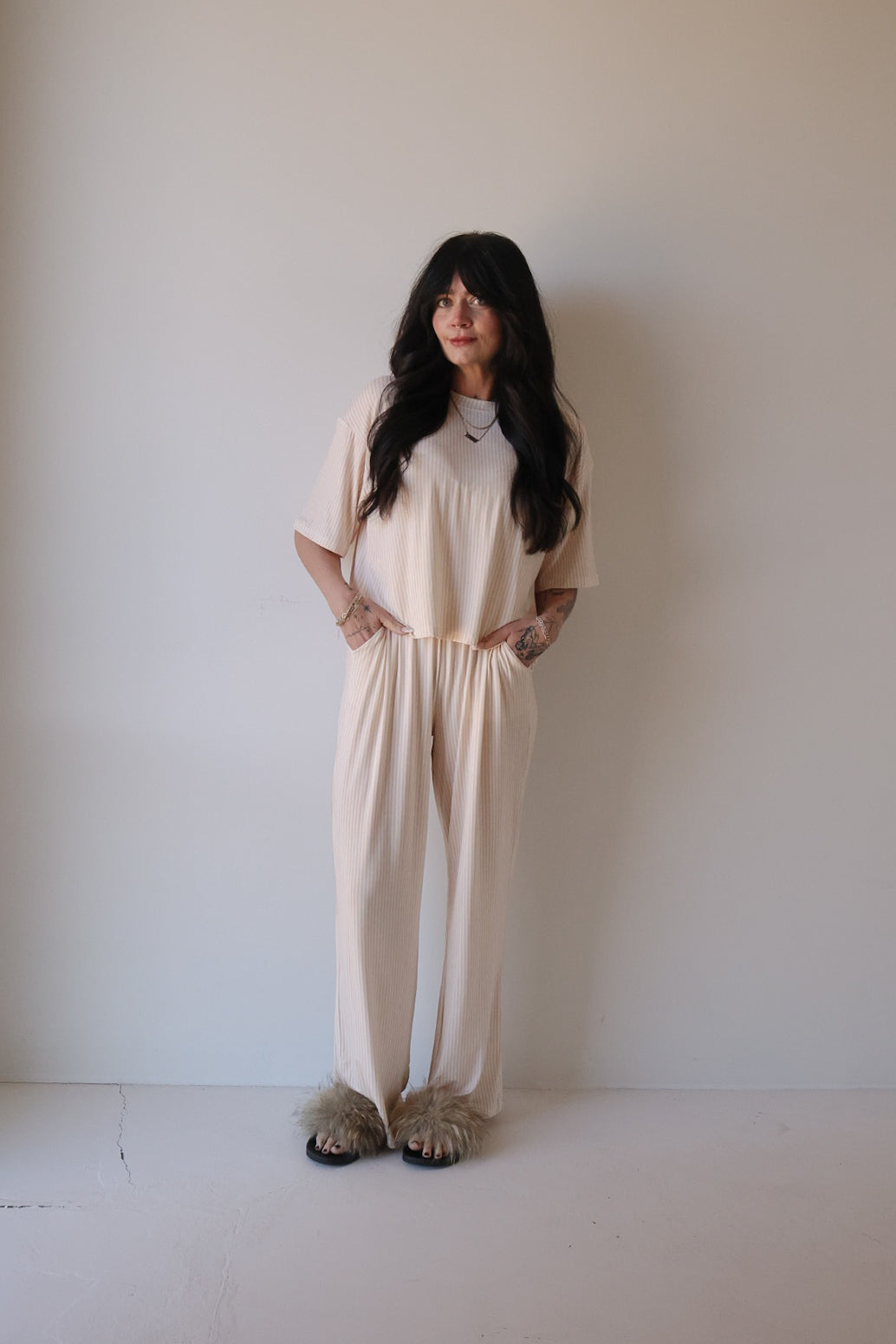 A woman with long dark hair stands against a plain wall, wearing the Women's Boxy Tee Lounge Set | Sadie Stripe from forever french baby and fluffy slippers. She smiles slightly with her hands in her pockets.