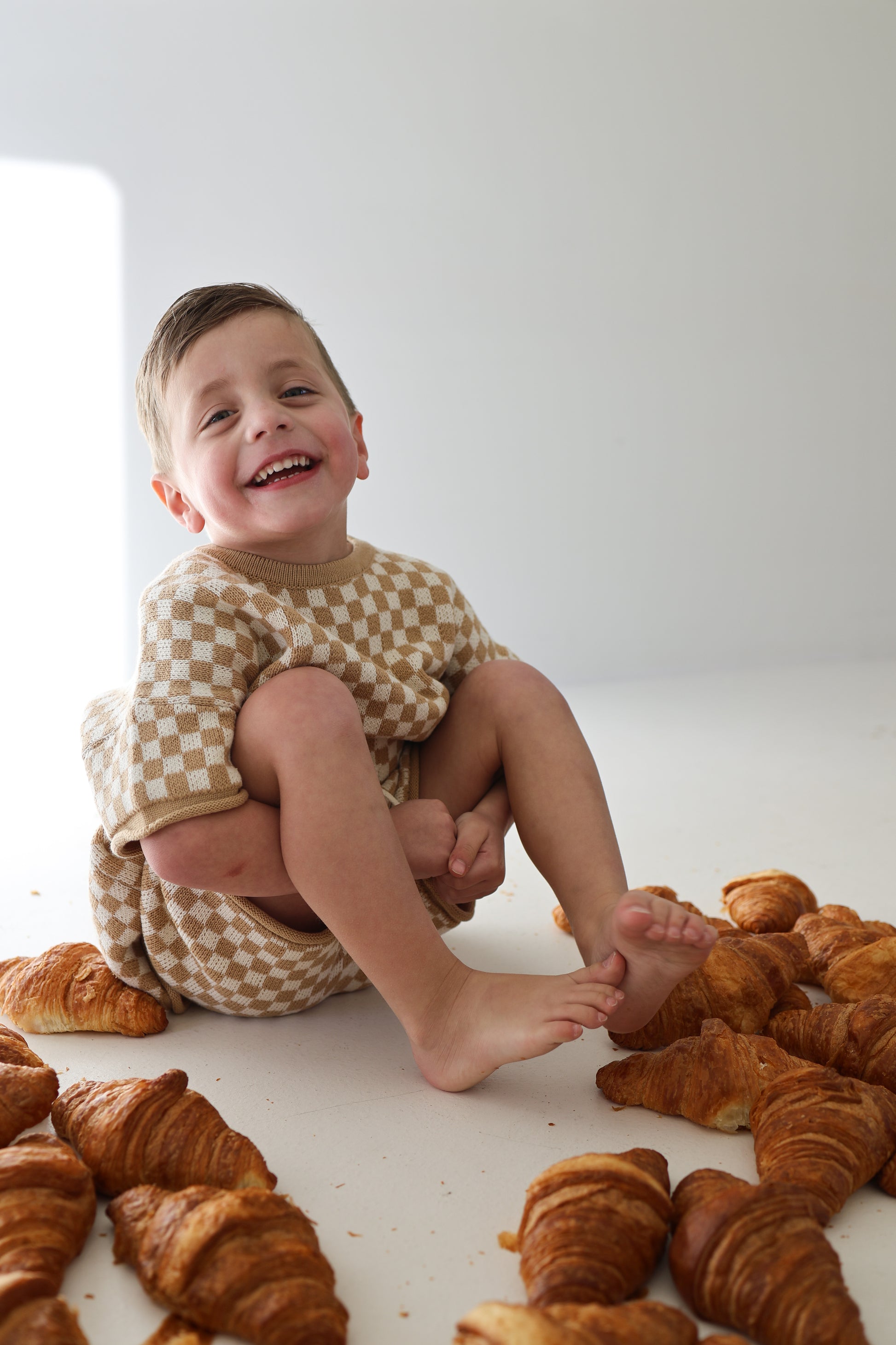 A smiling young child in the forever french baby Knit Short Set | Latte Check sits barefoot among croissants on a white floor, supported from behind by an adult’s arms.