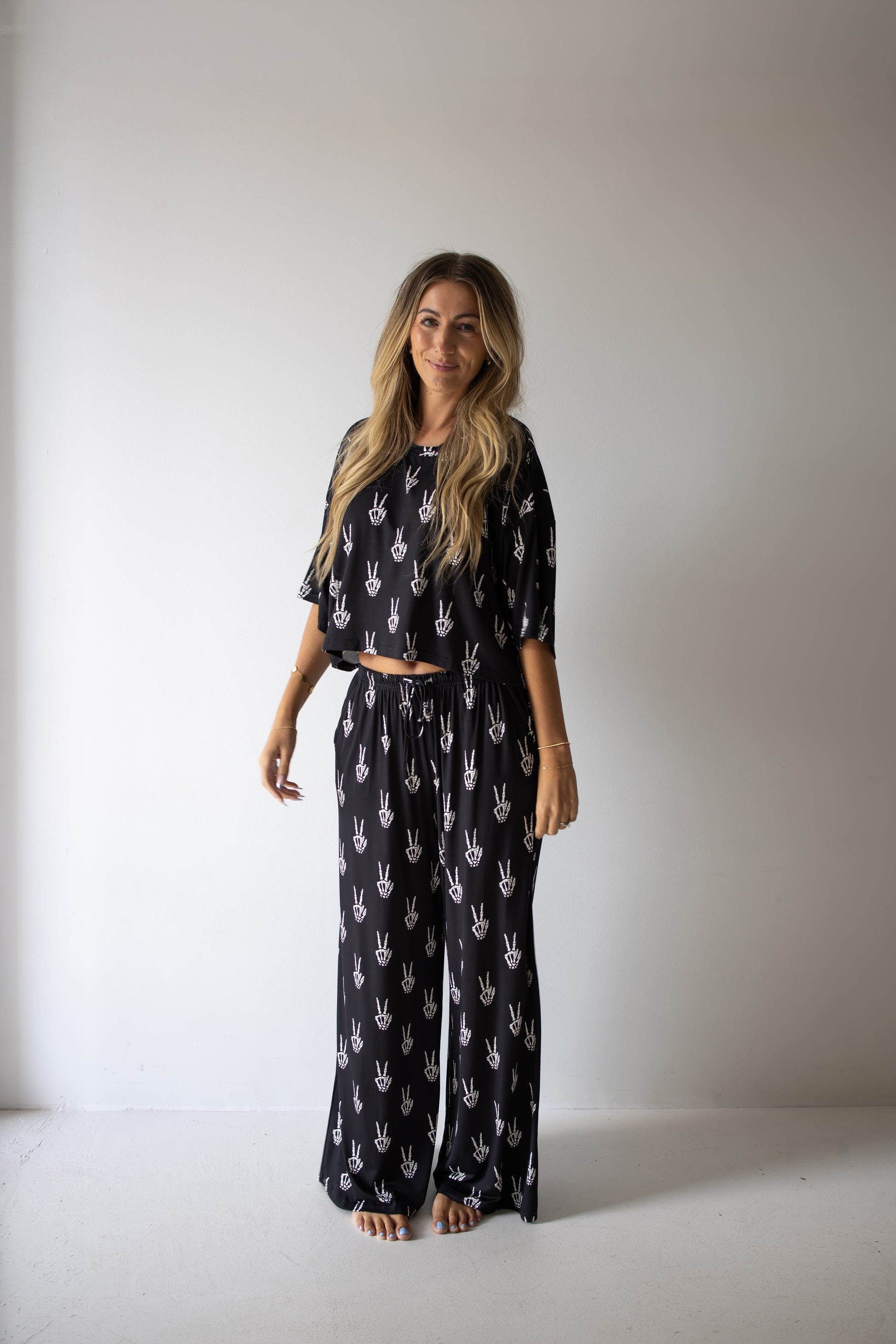A woman with long blonde hair stands barefoot against a plain white wall, smiling at the camera with her arms relaxed at her sides. She is wearing the forever french baby Women’s Bamboo Crop Tee Lounge Set in Midnight Skelly—a matching black crop tee and wide-leg pant set featuring a white hand print pattern.