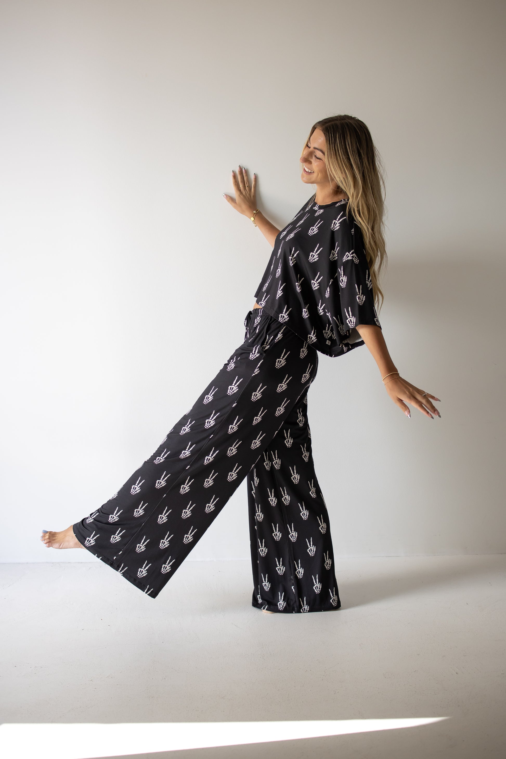 Wearing the Women’s Bamboo Crop Tee Lounge Set in Midnight Skelly by forever french baby, a woman poses playfully against a white wall, smiling with one leg raised and arms outstretched. The matching black outfit features a bamboo crop tee adorned with a white handprint pattern and wide-leg pants crafted from soft bamboo viscose.