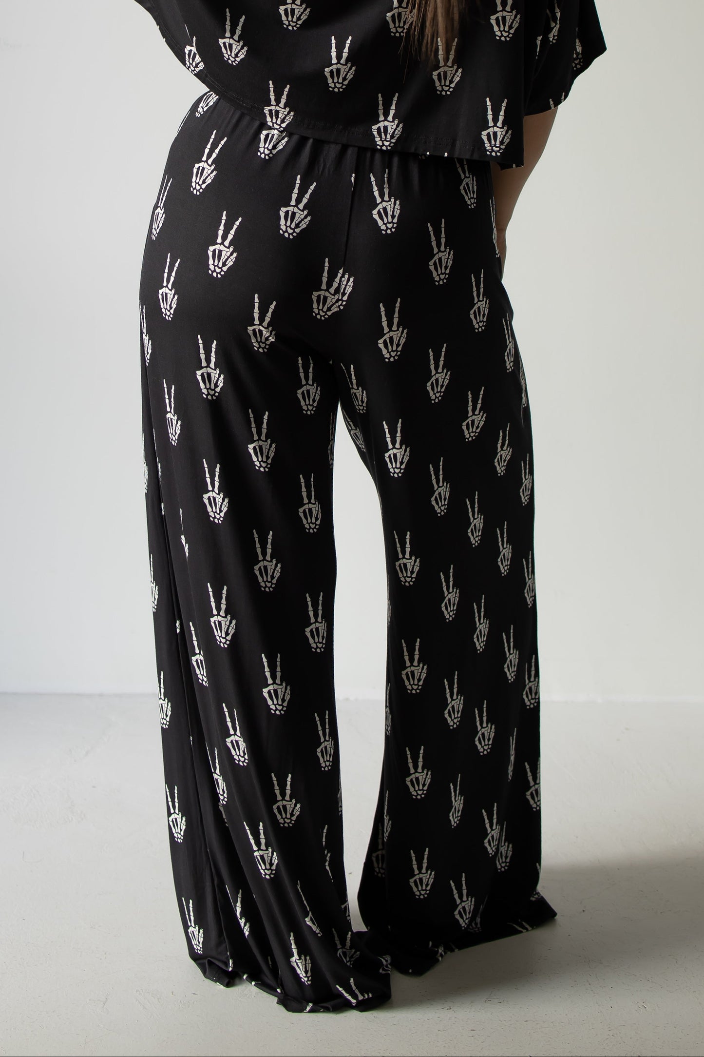 A person is seen from behind, standing against a plain light background, wearing the forever french baby Women’s Bamboo Crop Tee Lounge Set in the Midnight Skelly design—featuring black wide-leg pants and a matching crop tee adorned with a white hand peace sign pattern.