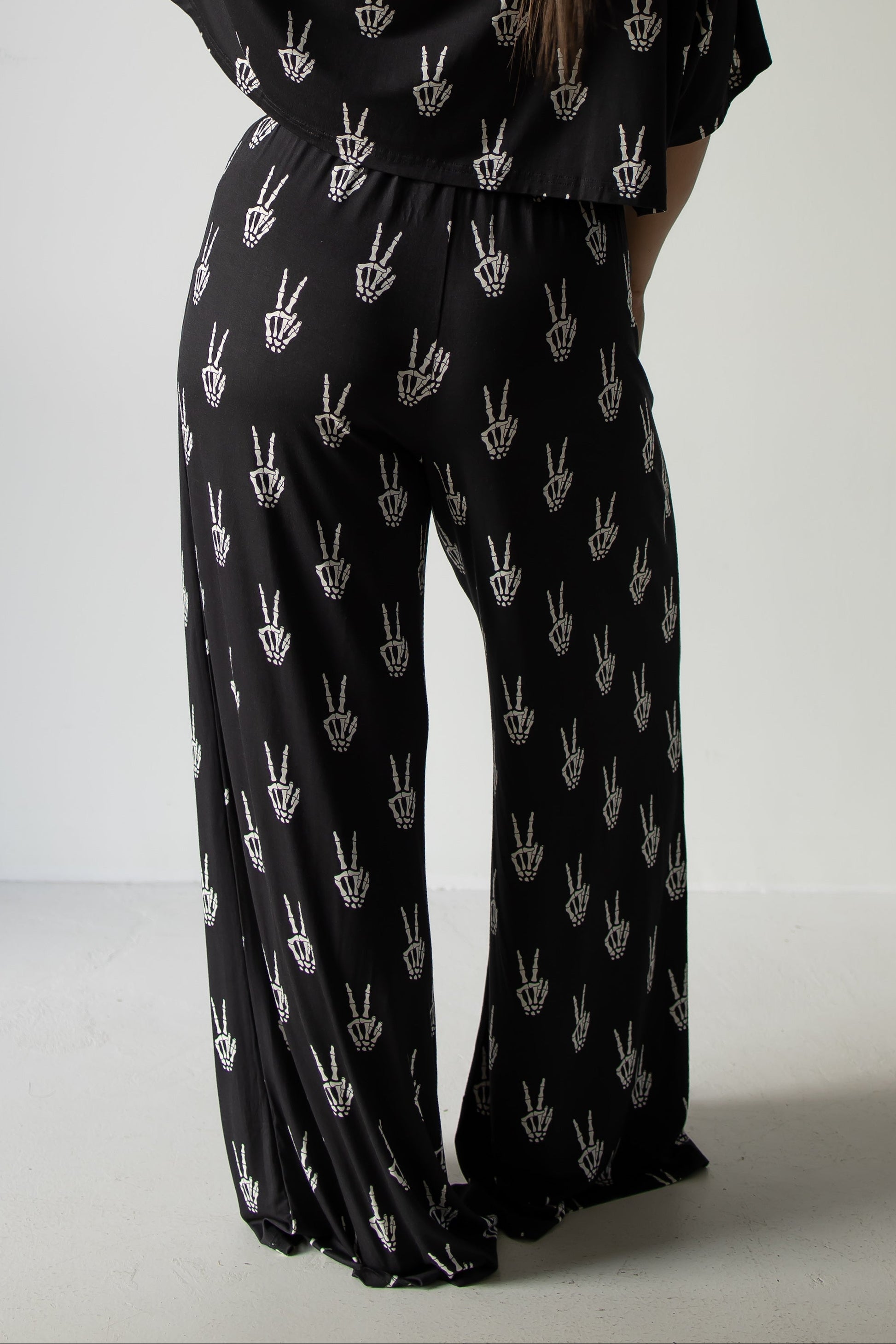 A person is seen from behind, standing against a plain light background, wearing the forever french baby Women’s Bamboo Crop Tee Lounge Set in the Midnight Skelly design—featuring black wide-leg pants and a matching crop tee adorned with a white hand peace sign pattern.