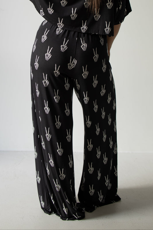 A person is seen from behind, standing against a plain light background, wearing the forever french baby Women’s Bamboo Crop Tee Lounge Set in the Midnight Skelly design—featuring black wide-leg pants and a matching crop tee adorned with a white hand peace sign pattern.