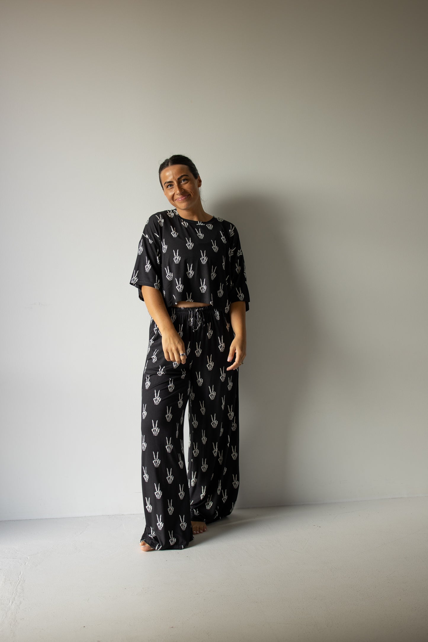 A person stands and smiles against a plain wall, wearing the forever french baby Women’s Bamboo Crop Tee Lounge Set in Midnight Skelly—a matching black set crafted from soft bamboo viscose with a white hand print pattern, featuring a loose crop tee and wide-leg pants.