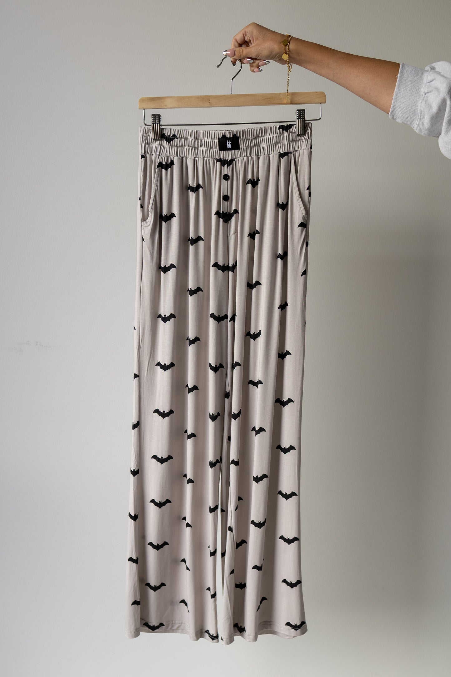 A hand holds a hanger displaying the forever french baby Short Sleeve Women’s Bamboo Lounge Set | It’s Bats!, featuring beige wide-leg pants with a black bat print against a plain background, showcasing the comfort and elegance of our hypo-allergenic bamboo clothing collection.