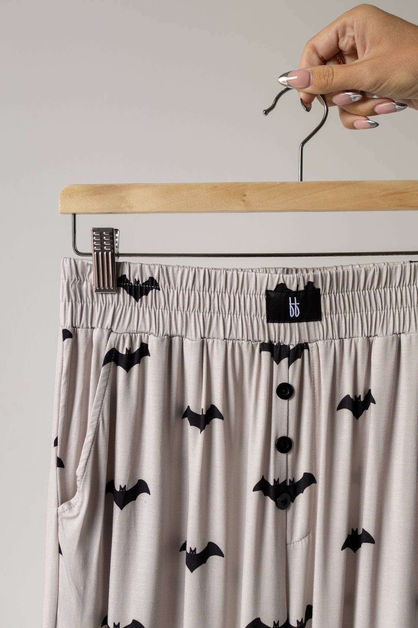 A hand holds a hanger with beige pajama pants, part of the forever french baby Short Sleeve Women’s Bamboo Lounge Set | It’s Bats!—a luxurious and hypo-allergenic bamboo set featuring a playful black bat print, elastic waistband, button-down front, and handy pockets for stylish comfort.