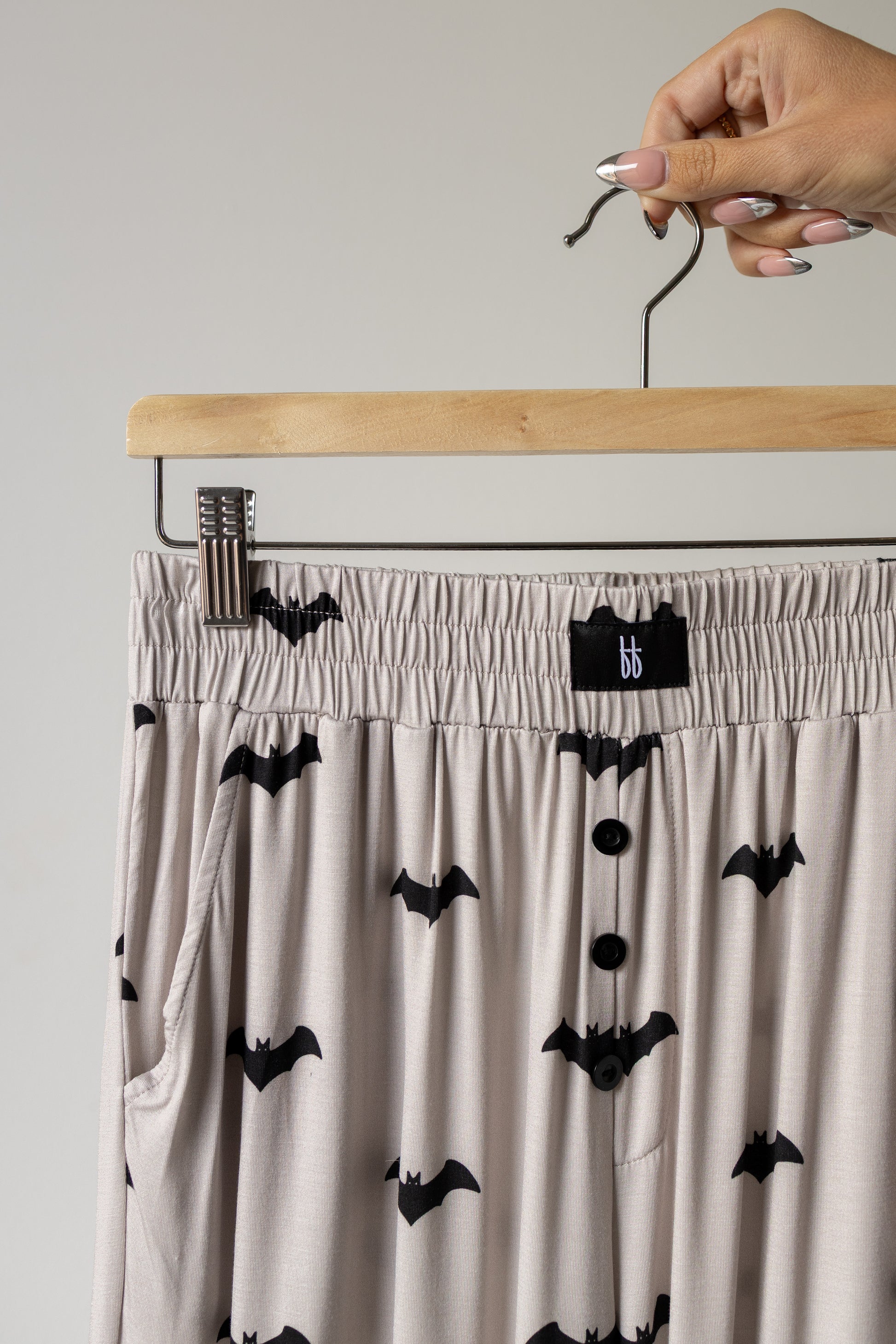 A hand holds a hanger with beige pajama pants, part of the forever french baby Short Sleeve Women’s Bamboo Lounge Set | It’s Bats!—a luxurious and hypo-allergenic bamboo set featuring a playful black bat print, elastic waistband, button-down front, and handy pockets for stylish comfort.