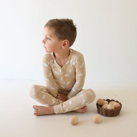 A young boy wears forever french baby’s Bamboo Two Piece Pajamas | Bunnies in beige, sitting cross-legged on the floor beside a small basket filled with wooden eggs and two eggs on the floor.