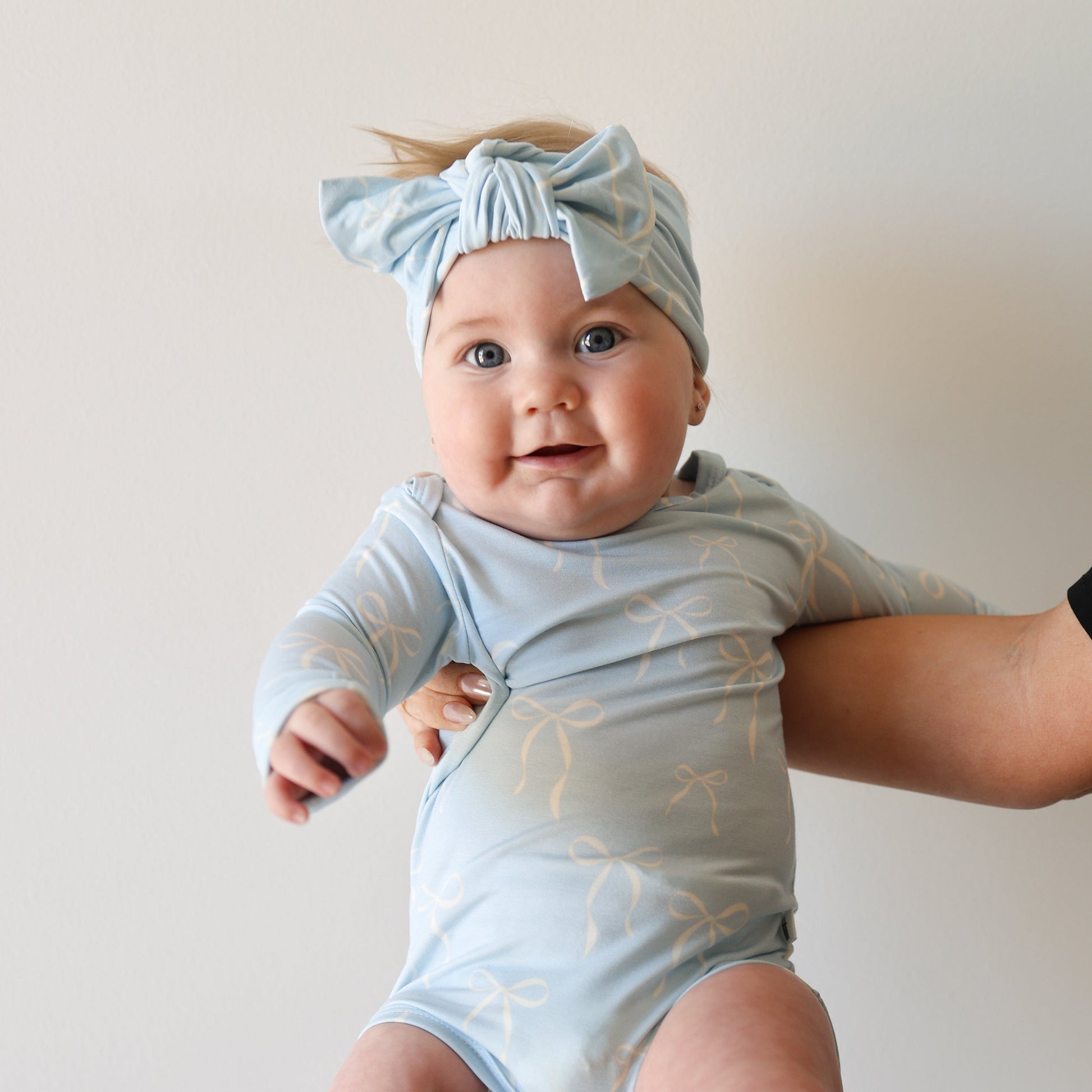 A baby in the forever french baby Bamboo Head Wrap | Blue Bows, paired with a bow-patterned long-sleeve onesie, is held by an adult's hand against a plain white background.