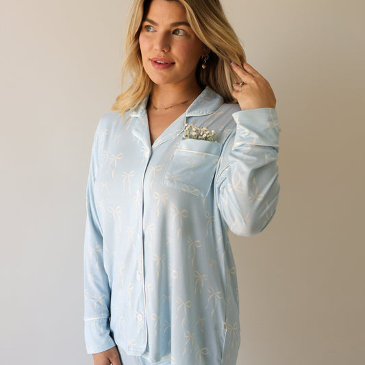 A woman with blonde hair wears the forever french baby Women's Bamboo Button Up Lounge Set | Blue Bows, standing against a plain background, touching her hair with white flowers tucked into her chest pocket.