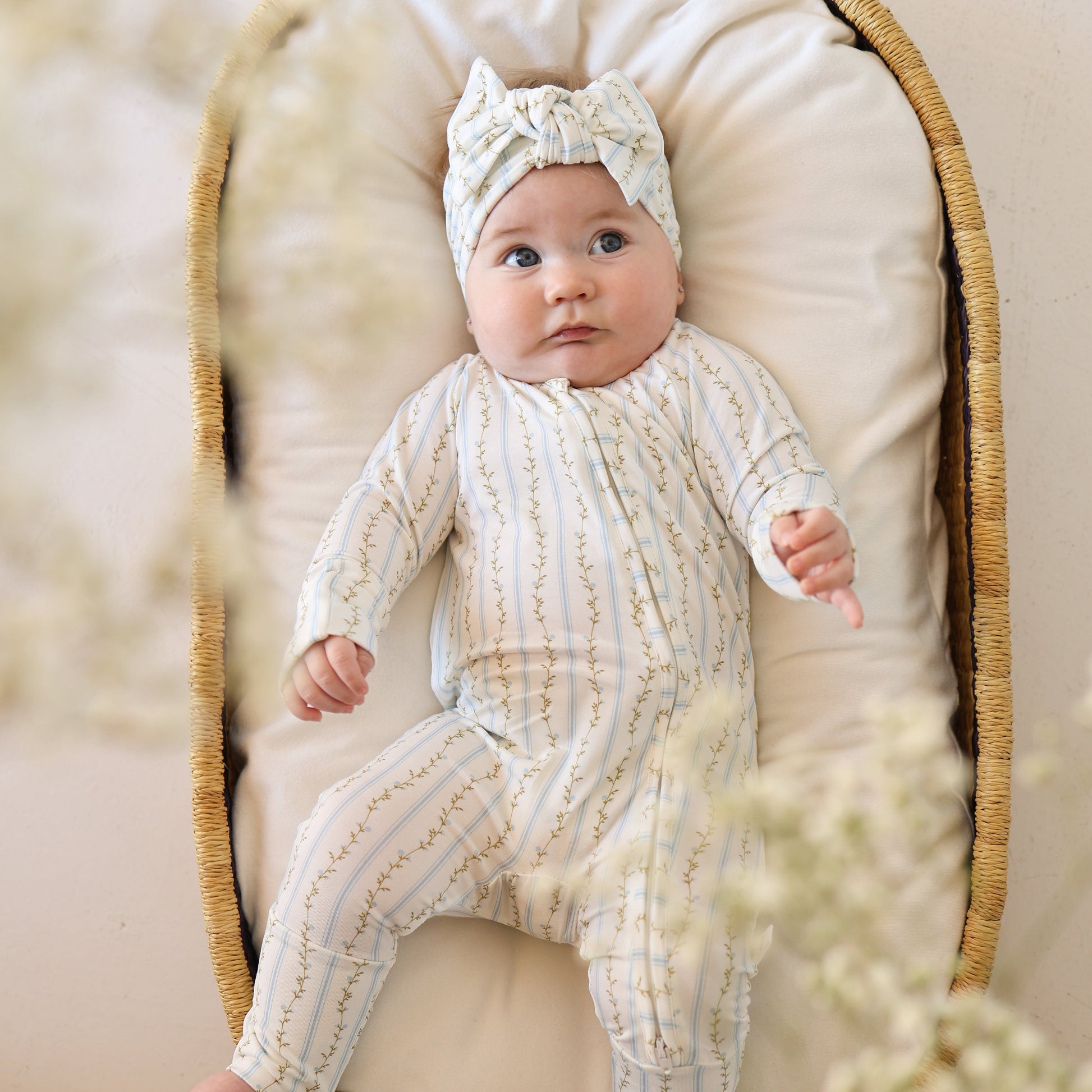 A baby in forever french baby's Bamboo Zip Pajamas | Dainty Vines lies on a cushioned basket, gazing up with wide eyes as soft, blurred flowers frame the foreground.