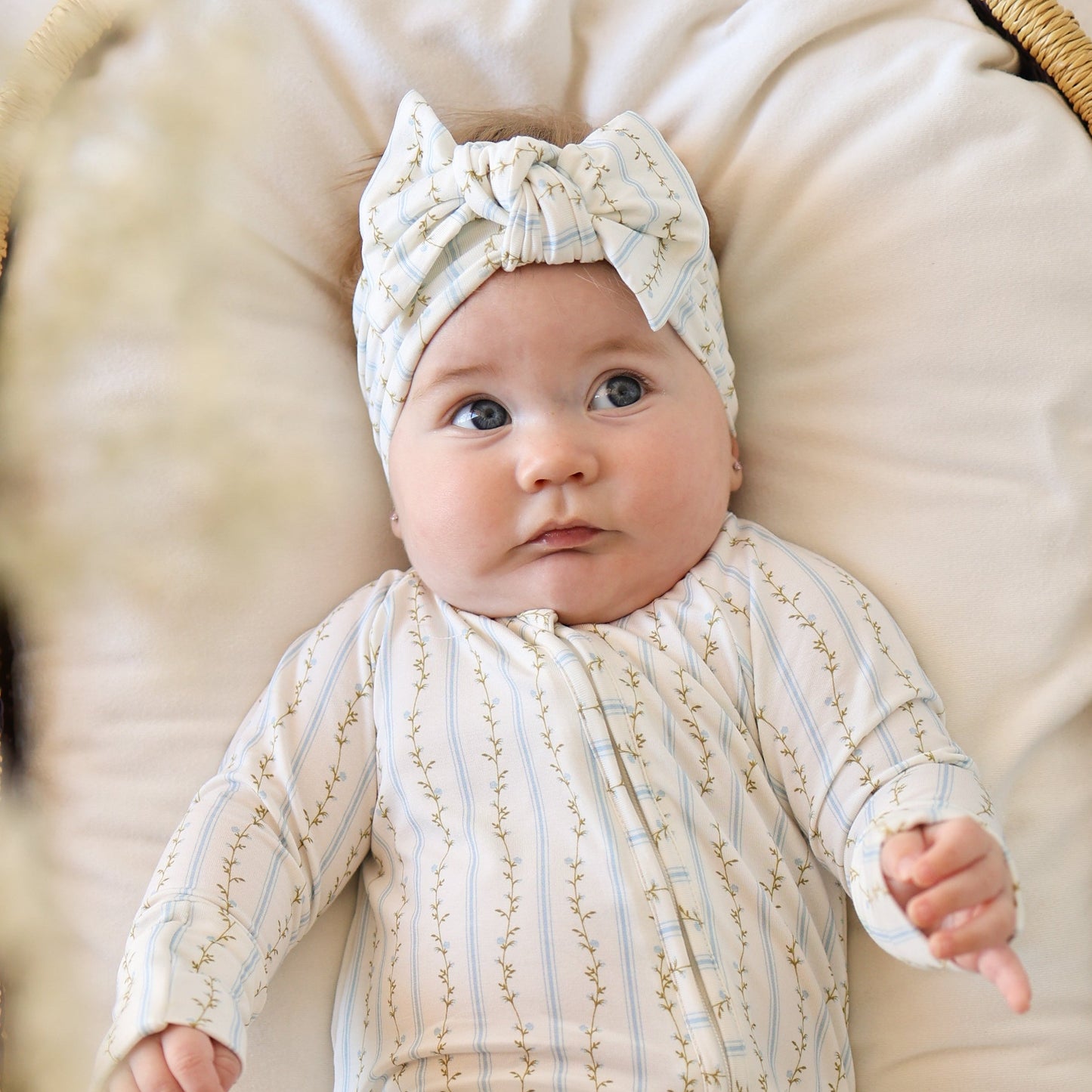 A baby in a matching light floral onesie and Forever French Baby Bamboo Head Wrap | Dainty Vines lies on a soft surface, gazing up with wide eyes and a curious look.