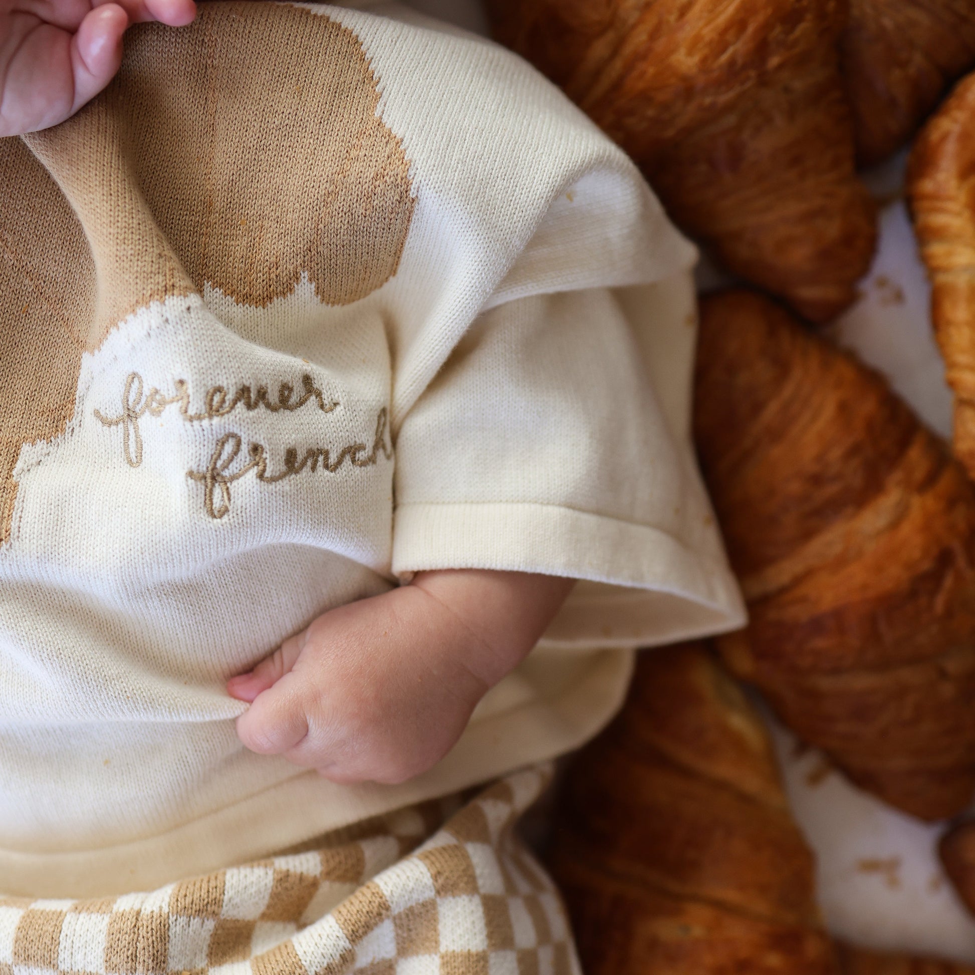 A baby wearing the forever french baby Knit Pant Set in Croissants lies next to a pile of croissants, wrapped in a checkered blanket. Only the baby's hand, arm, and part of the face are visible.