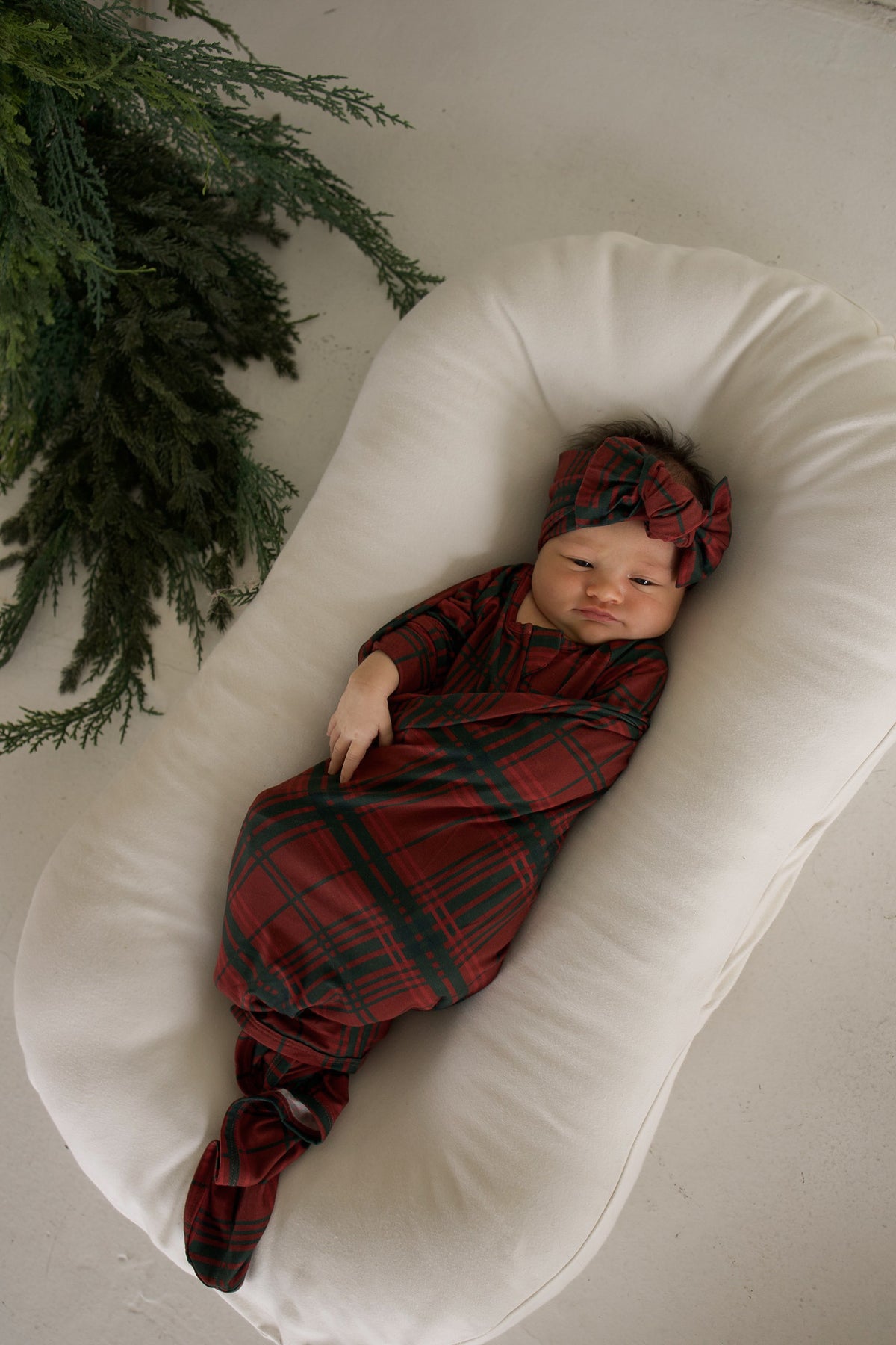 A newborn baby wearing a Bamboo Swaddle | Christmas Plaid and matching headwrap, resting in a cream-colored lounger.