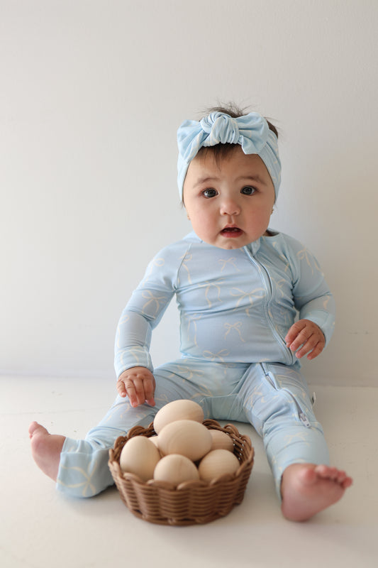 A baby in forever french baby's Bamboo Zip Pajamas | Blue Bows with a matching headband sits on the floor next to a wicker basket of wooden eggs, against a plain white background.