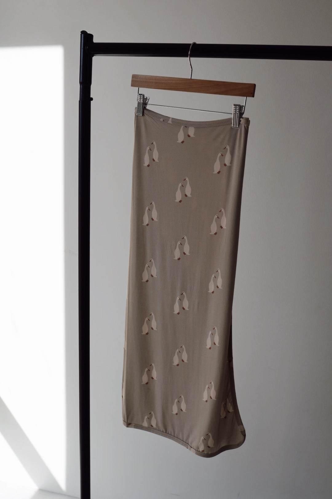 A Bamboo Swaddle by forever french baby, called "Silly Goose," hangs gracefully on a wooden hanger from a black rack against a white wall, catching natural light and casting gentle shadows nearby.