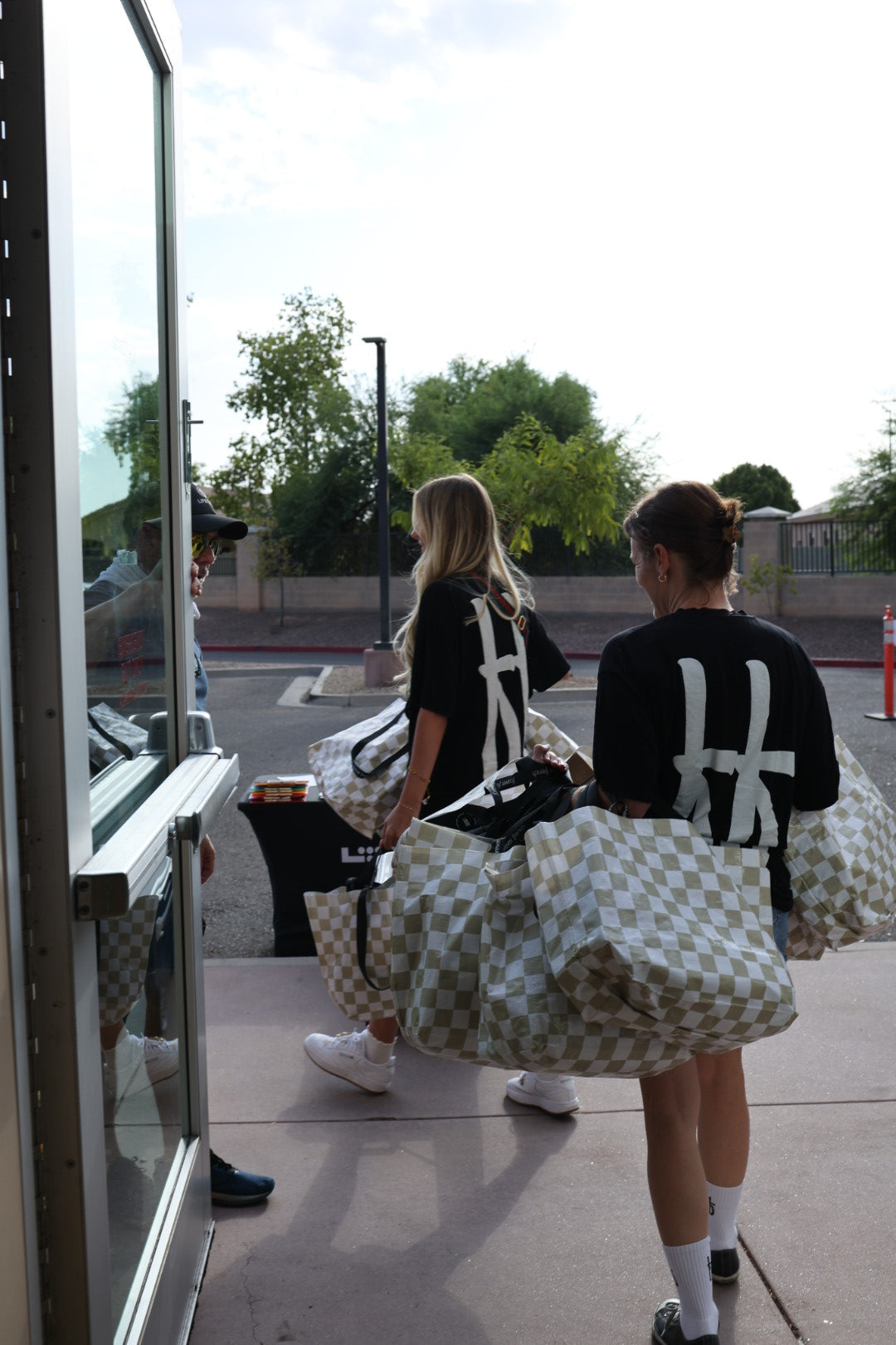 Two people in matching black shirts with white designs carry ff Warehouse Sale Totes from forever french baby and walk toward an open glass door, where another person waits outside on a sunny day. Trees and a fence are visible in the background.