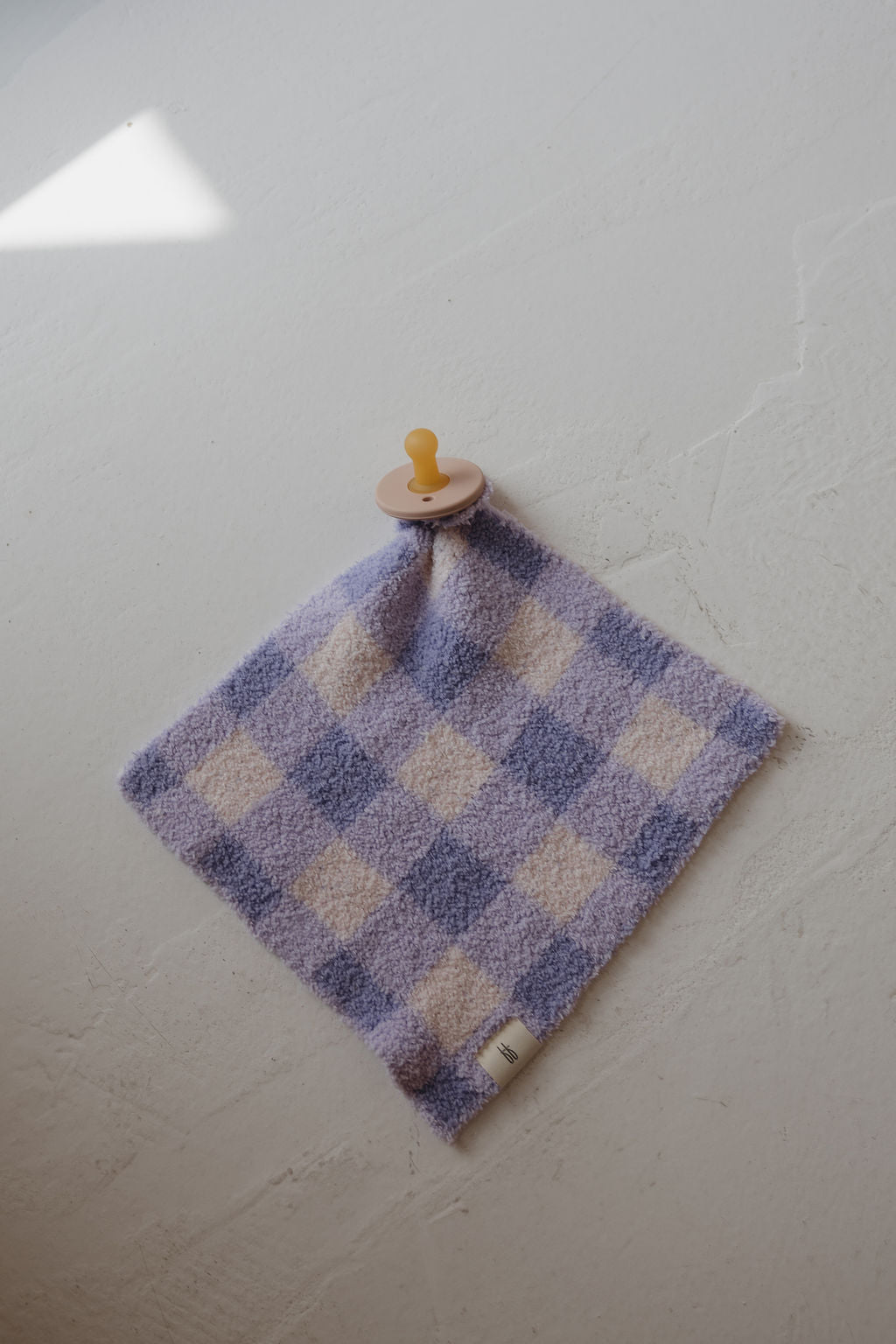 A yellow pacifier is attached to the corner of the Lovey | Marilyn microfiber baby blanket by forever french baby, featuring a soft, checkered purple and white design and hanging on a textured white wall with sunlight in the top left corner.