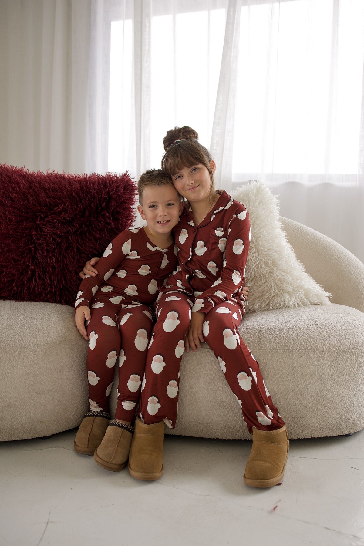 A boy and a girl sit together on a sofa wearing matching Bamboo Two Piece Pajamas | Vintage Santa with a red and white Santa Claus pattern.