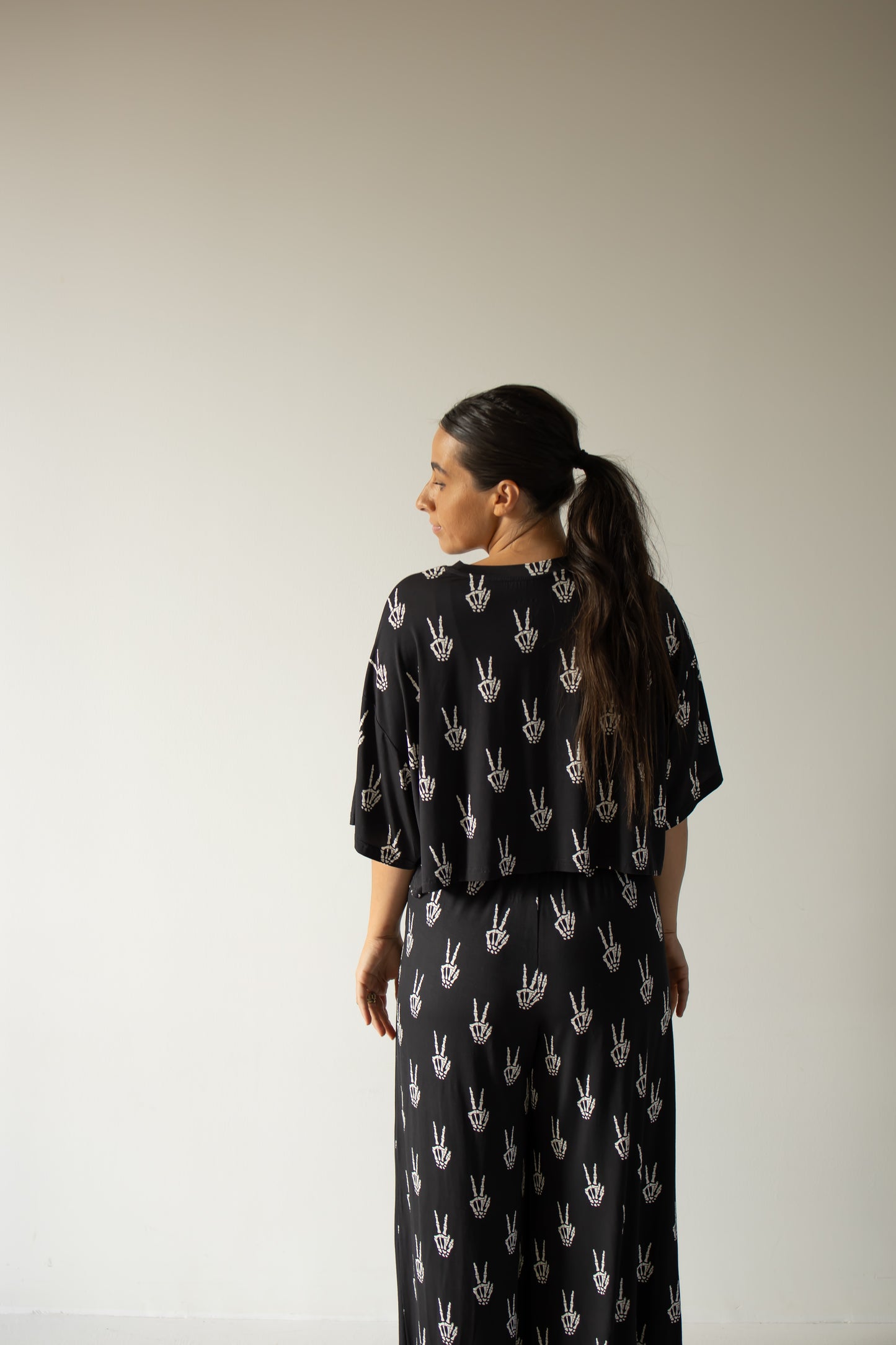 A woman with long dark hair in a ponytail stands facing away against a plain light-colored background, wearing the forever french baby Women’s Bamboo Crop Tee Lounge Set in Midnight Skelly, featuring a black design with a white hand print pattern crafted from soft bamboo viscose.