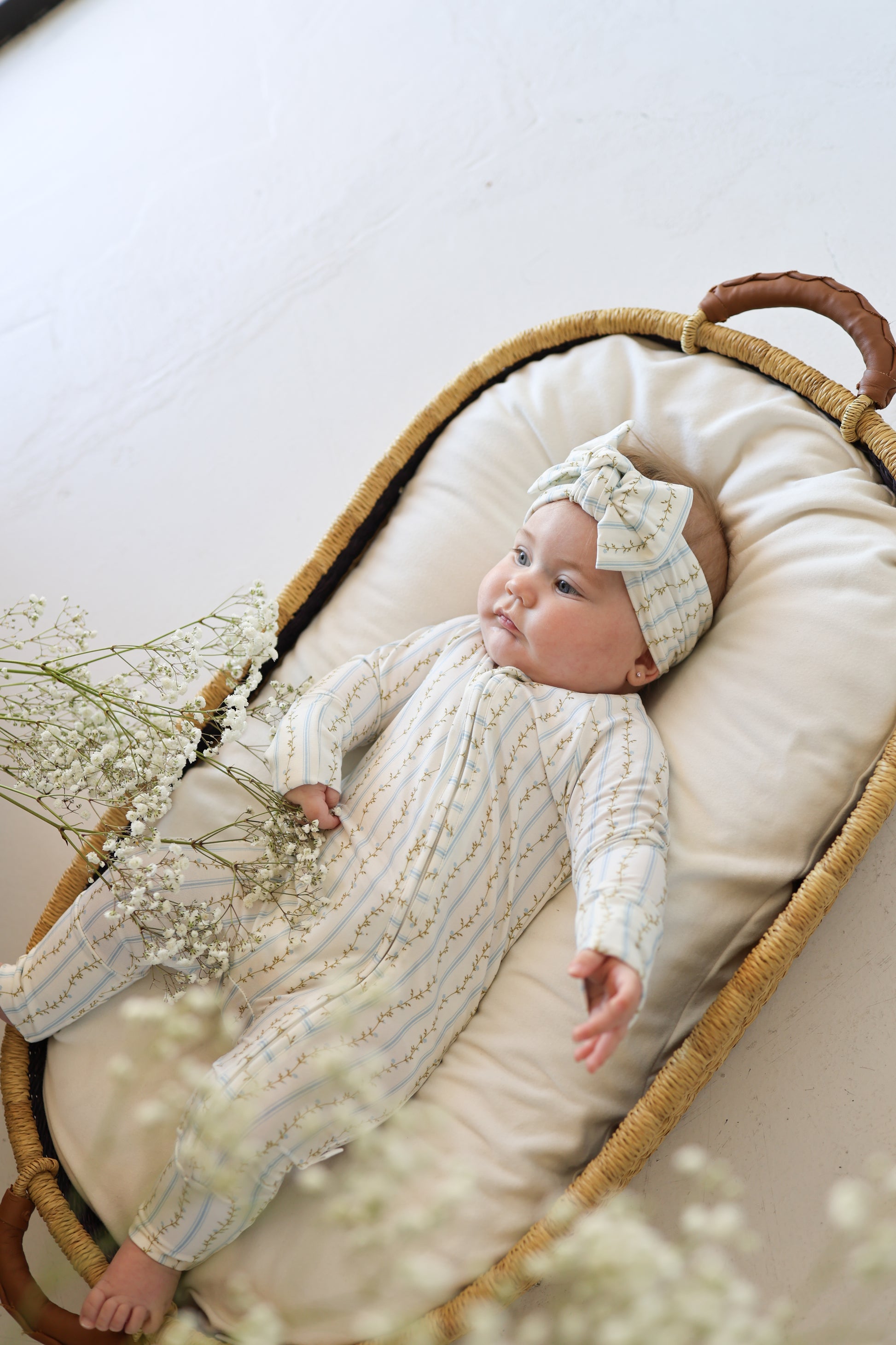 A baby in forever french baby's Bamboo Zip Pajamas | Dainty Vines with a matching headband lies calmly in a woven basket on a soft surface, holding baby's breath flowers and gazing upward.