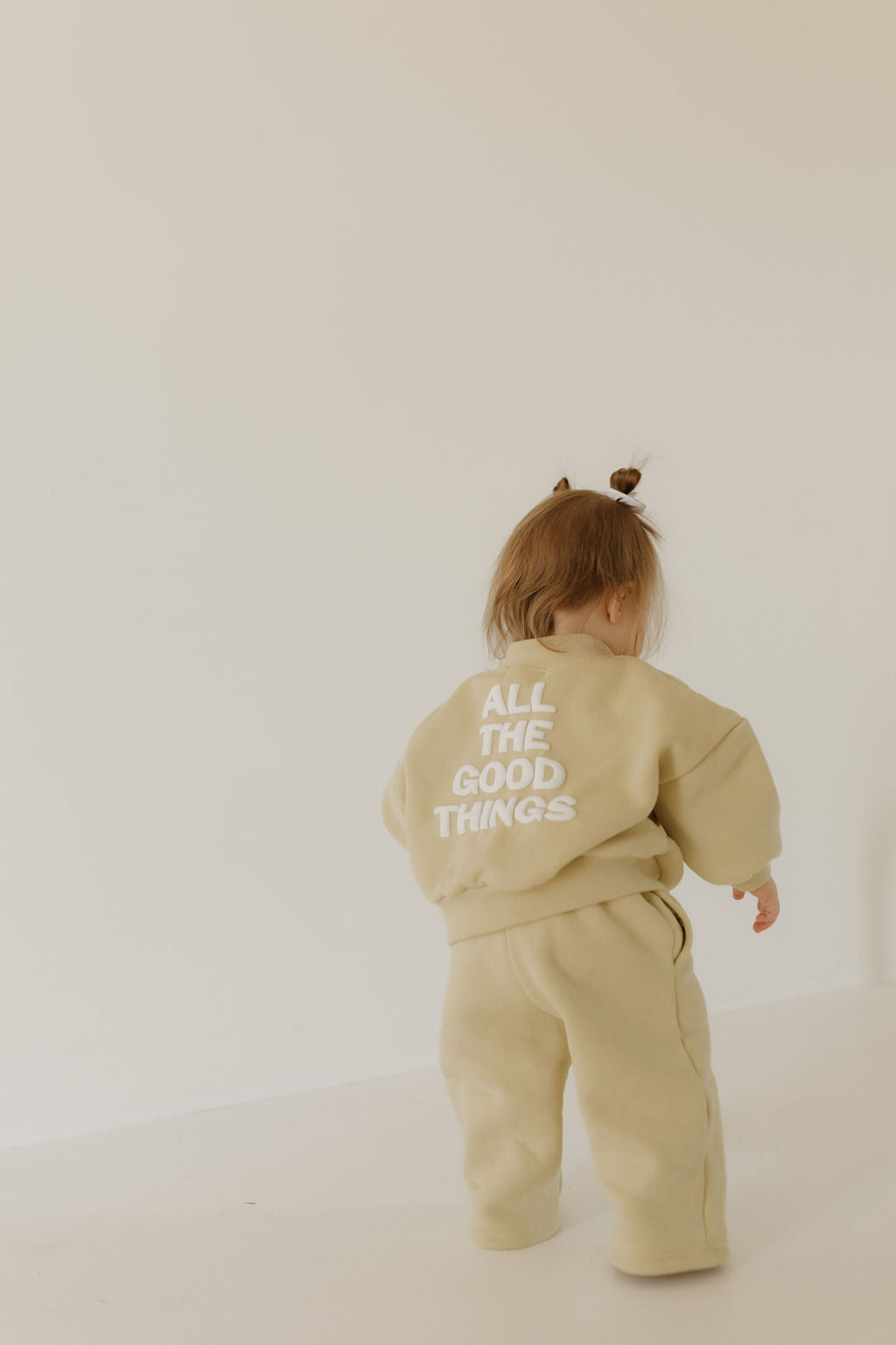 A toddler with short brown hair, styled in two small buns, is dressed in the "All the Good Things" sweat set by forever french baby. The child stands against an off-white background, showcasing a timeless moment of youthful style.