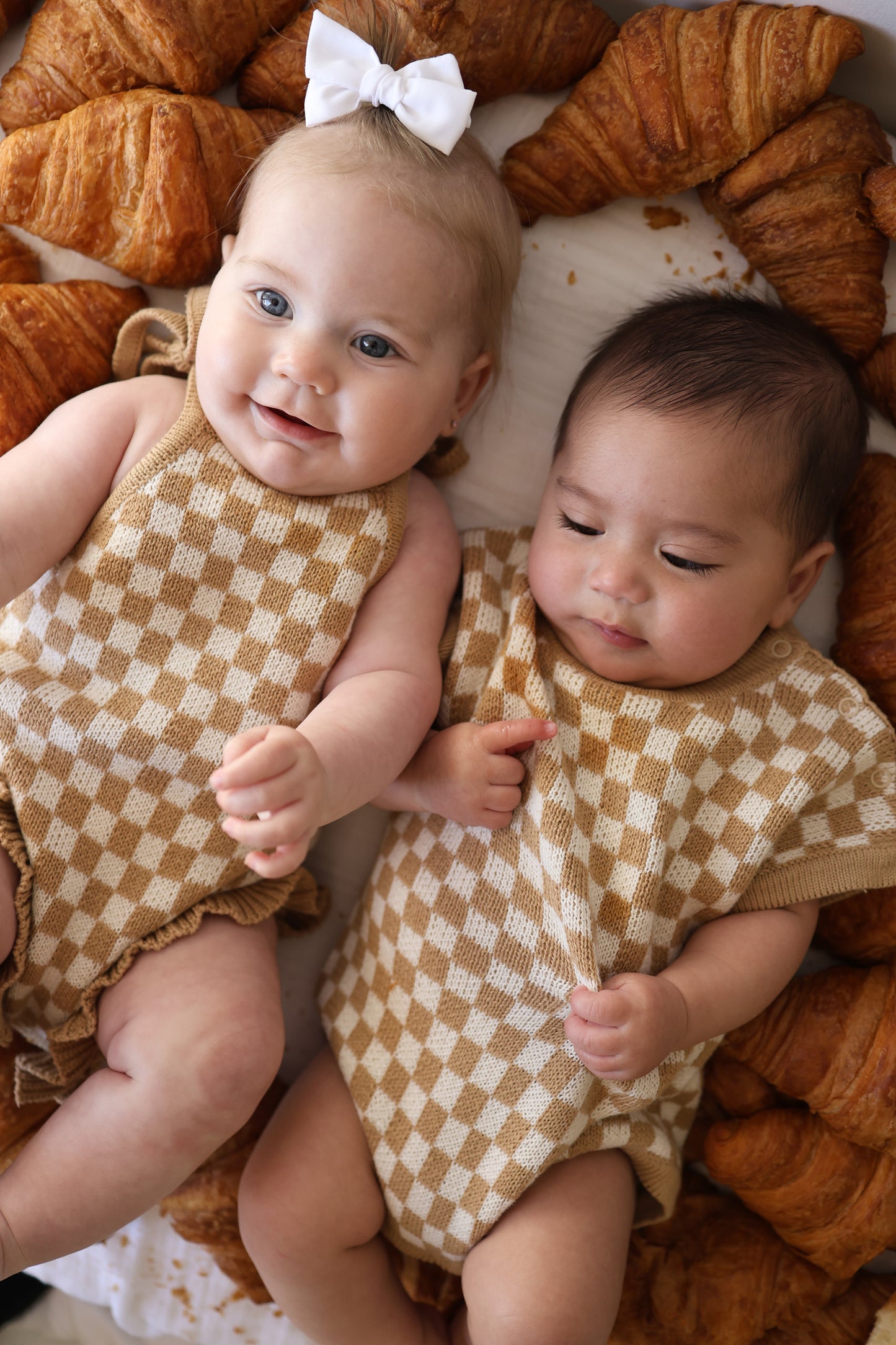 Two babies wearing forever french baby Knit Romper | Latte Check lie side by side on a bed with croissants—one in a white bow with blond hair and light skin, the other with darker hair and medium skin.