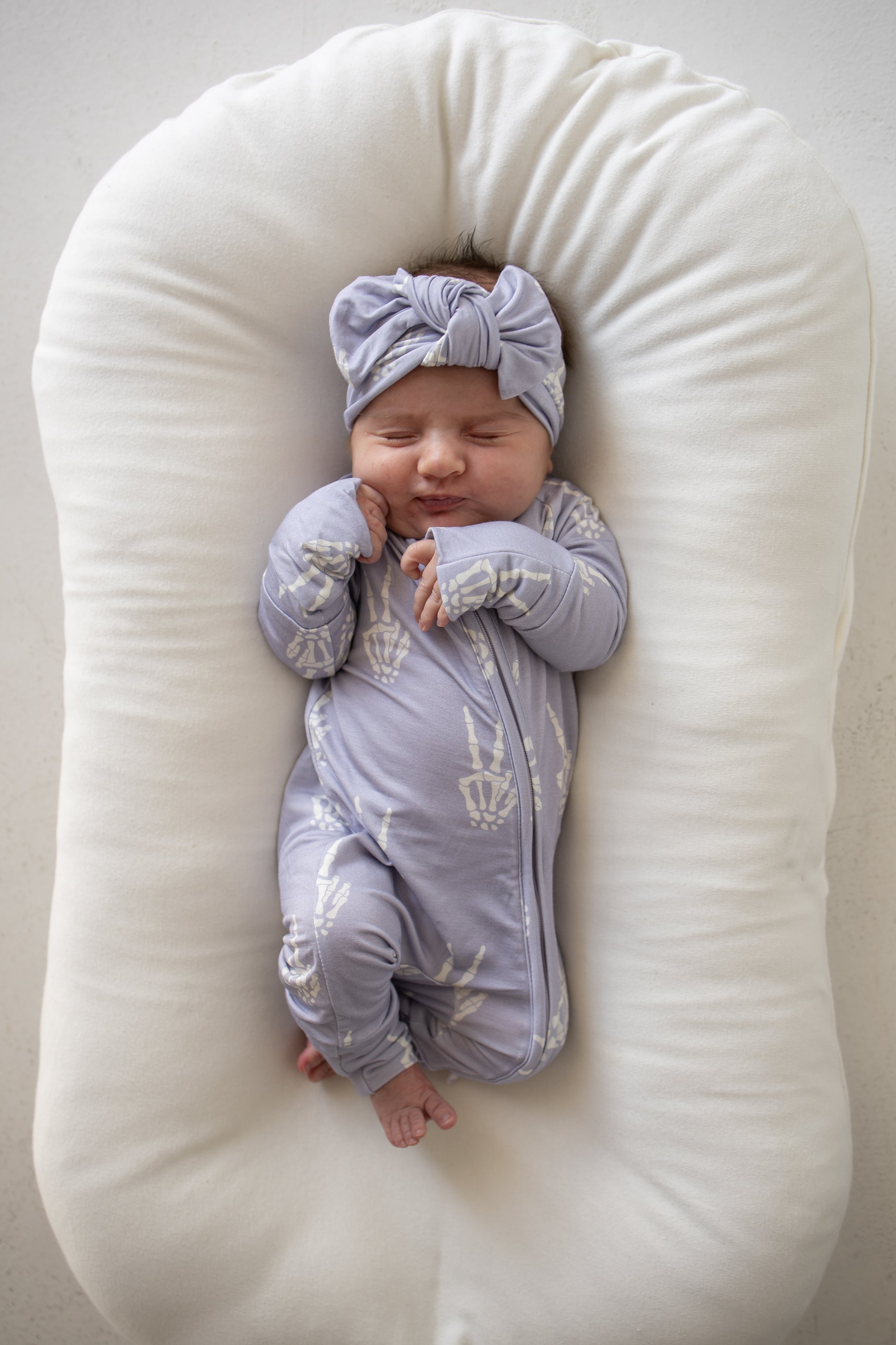 A baby wearing forever french baby’s Bamboo Zip Pajamas in the Enchanted Skelly print and a matching headband sleeps on a white, cushioned lounger.