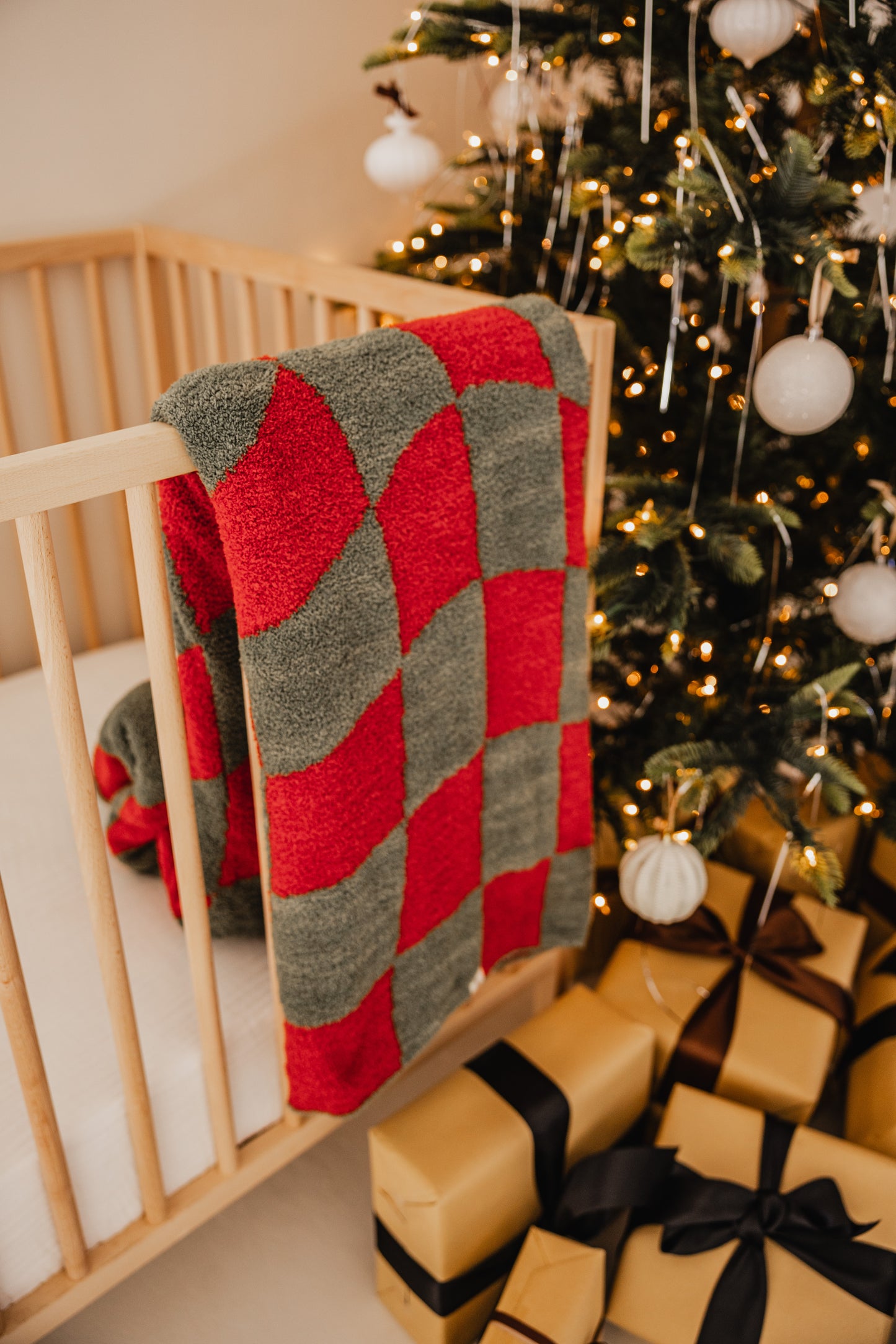 A plush Retro Christmas blanket from forever french baby, crafted from microfiber feather yarn, drapes over a wooden crib next to a tree adorned with lights and white ornaments. Neatly wrapped gold gifts with black ribbons sit beneath the tree, enhancing the festive nostalgia.