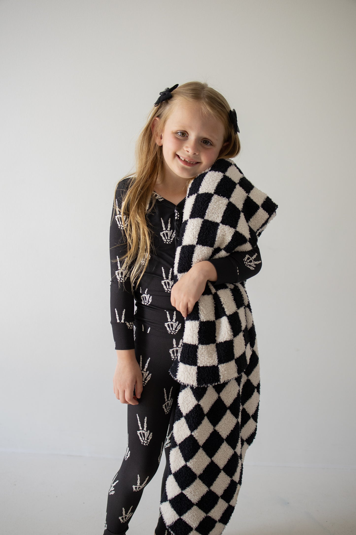 A young girl with long blonde hair, wearing the forever french baby Bamboo Two Piece Pajamas in the Midnight Skelly print, smiles while holding a black and white checkered blanket against a plain light background.