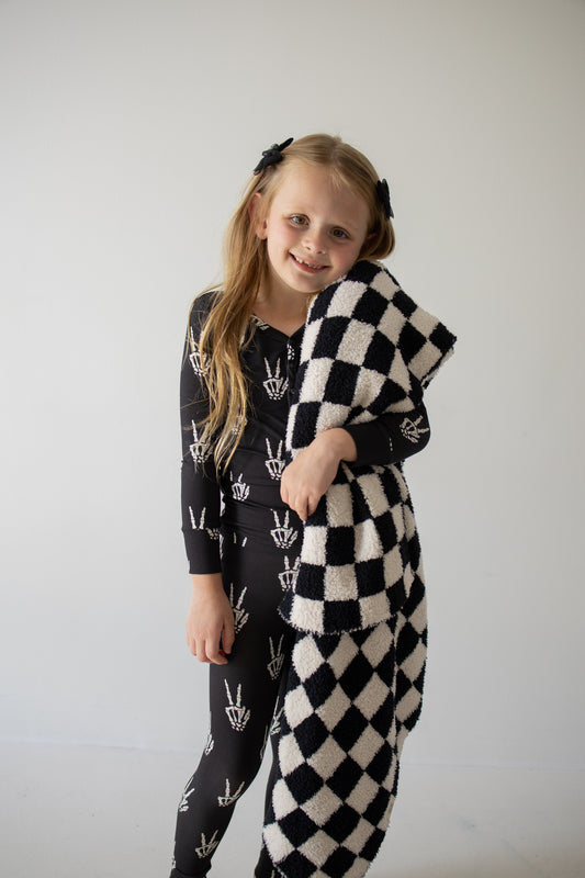 A young girl with long blonde hair, wearing the forever french baby Bamboo Two Piece Pajamas in the Midnight Skelly print, smiles while holding a black and white checkered blanket against a plain light background.