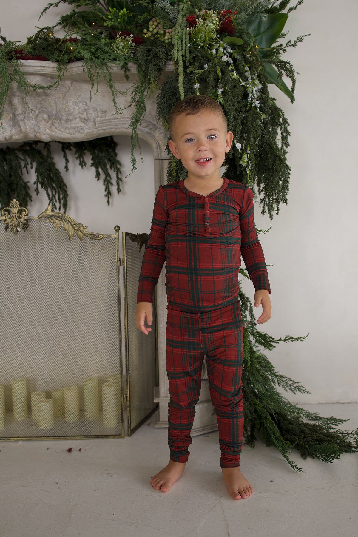A young boy stands in front of a fireplace wearing Bamboo Two Piece Pajamas | Christmas Plaid.