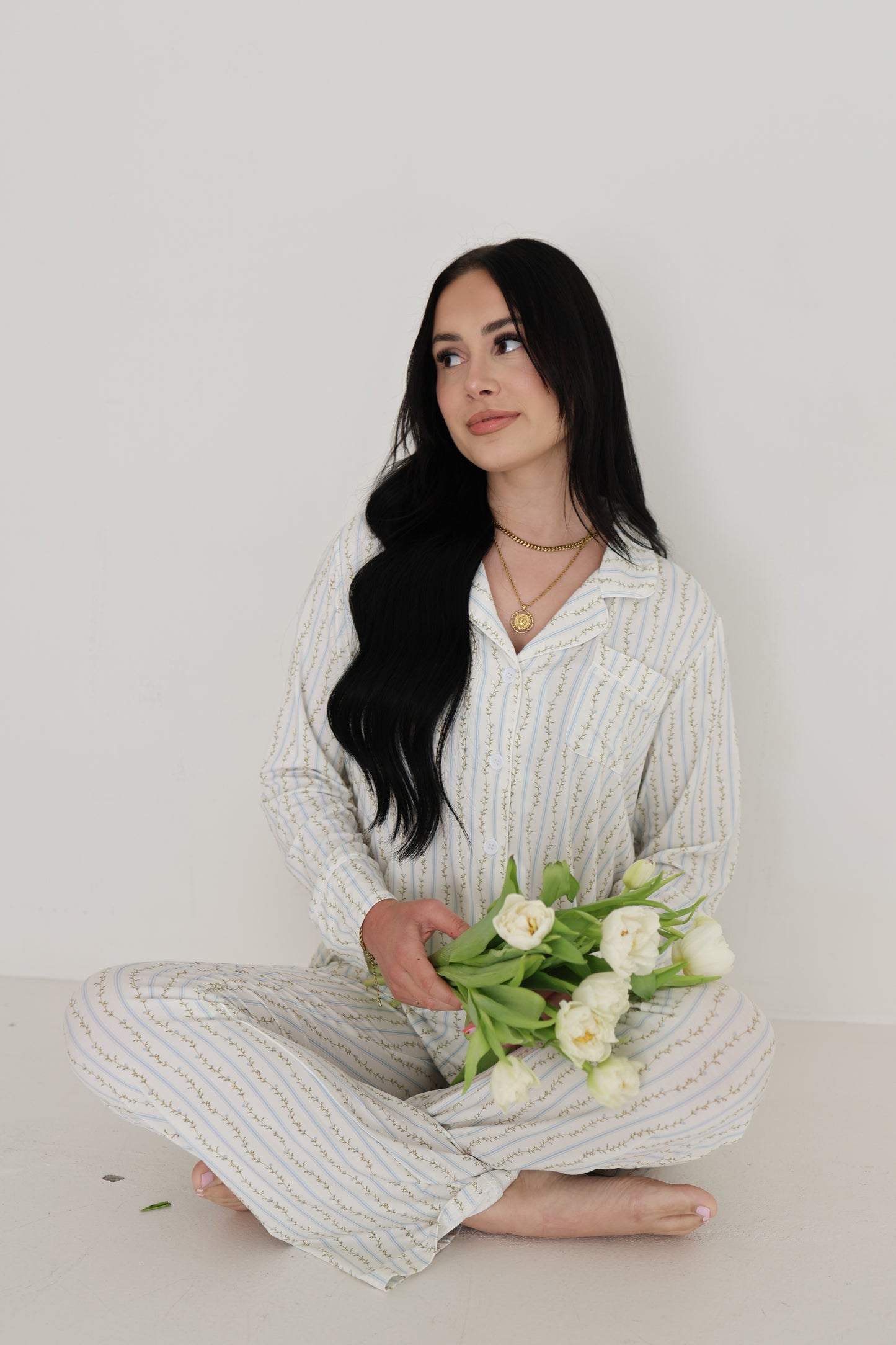 A woman with long dark hair sits cross-legged in the forever french baby Women's Bamboo Button Up Lounge Set | Dainty Vines, holding white tulips and gazing thoughtfully to the side against a plain white background.