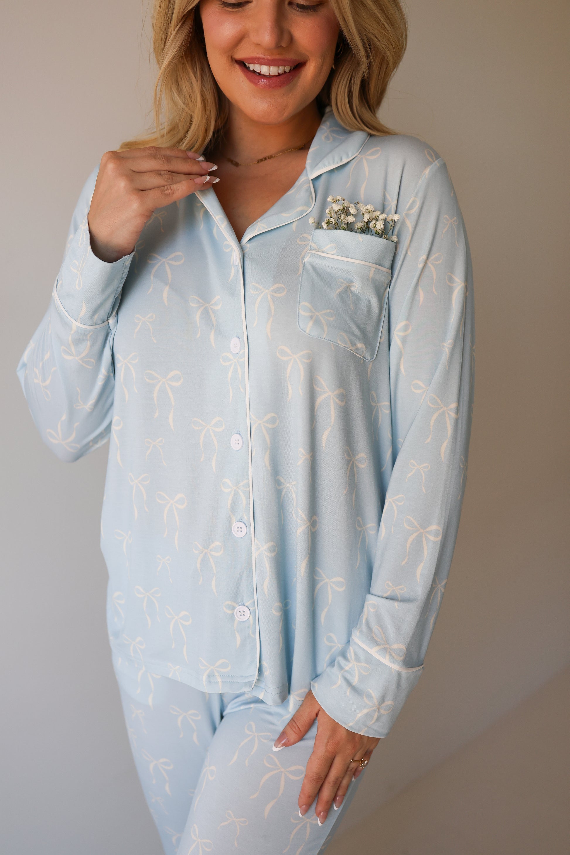 A woman in the forever french baby Women's Bamboo Button Up Lounge Set | Blue Bows smiles, showing her teeth. A small sprig of white flowers is tucked into the chest pocket of her blue pajama top with a white bow pattern.