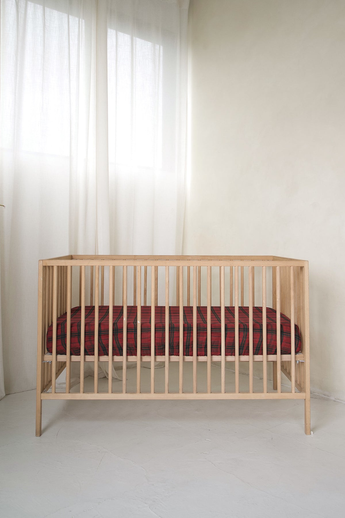 A wooden crib fitted with a Muslin Crib Sheet | Christmas Plaid in a red and black pattern.