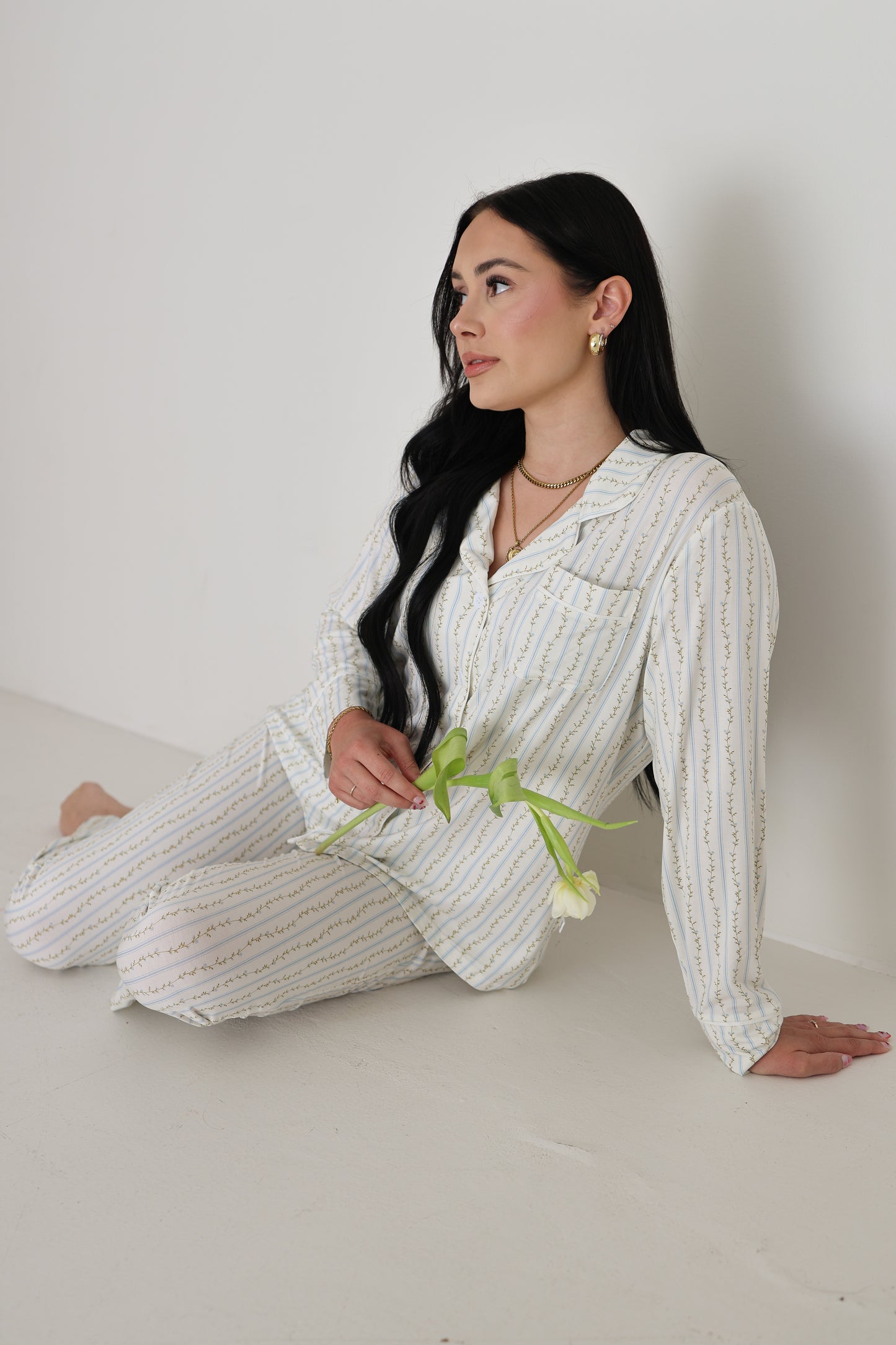 A woman with long dark hair sits on the floor in a forever french baby Women's Bamboo Button Up Lounge Set | Dainty Vines, holding a white tulip and enjoying the comfort of this hypo-allergenic bamboo loungewear.